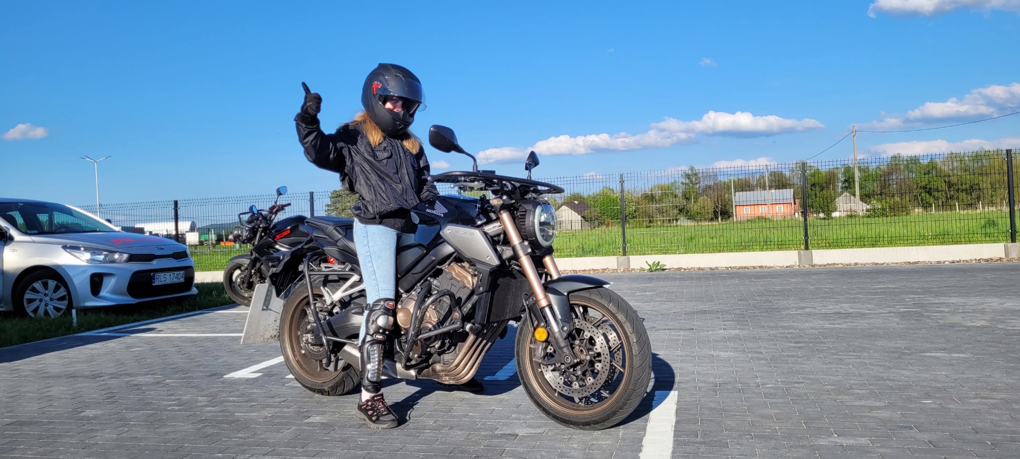 Kobieta w kasku na motocyklu Honda CB650R, uniesiona ręka w geście aprobaty. W tle zaparkowane motocykle i samochód, błękitne niebo.