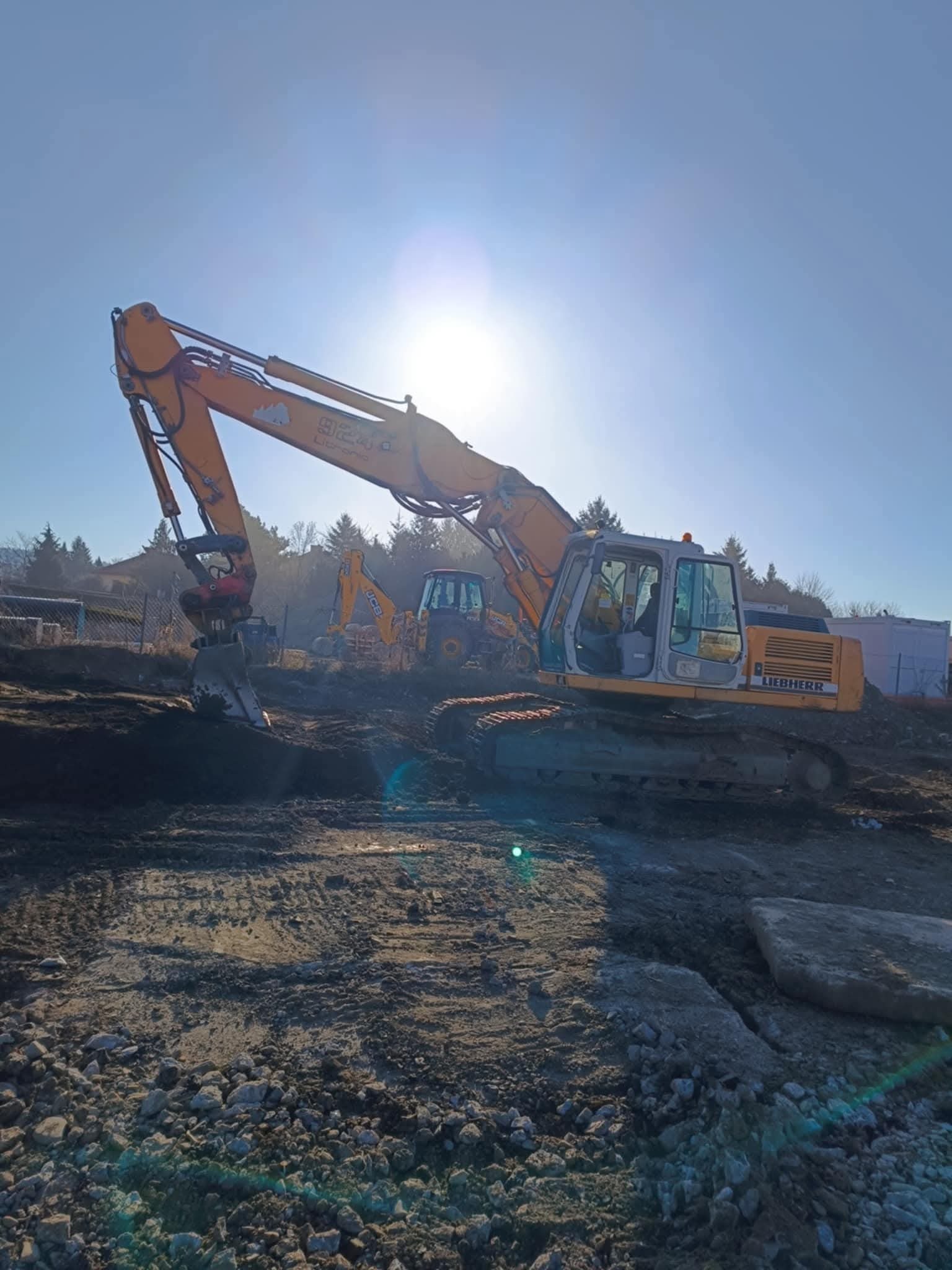 Żółta koparka Liebherr na placu budowy w słoneczny dzień, z widoczną drugą koparką w tle. Prace ziemne w toku, teren przygotowany pod budowę.