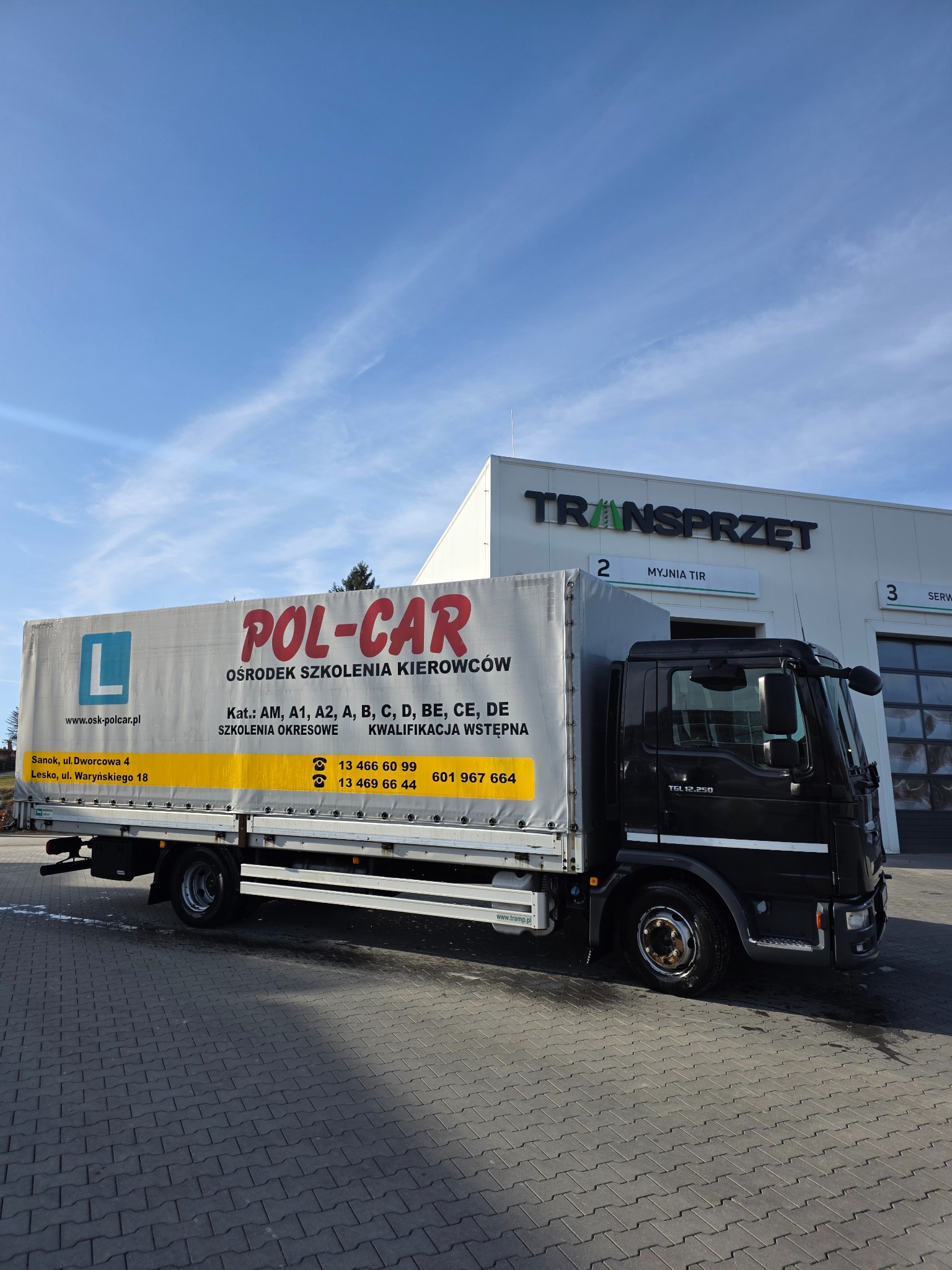 Ciężarówka POL-CAR, ośrodka szkolenia kierowców, z oznaczeniem 'L' i numerami kontaktowymi, zaparkowana przed budynkiem Transprzęt w słoneczny dzień.