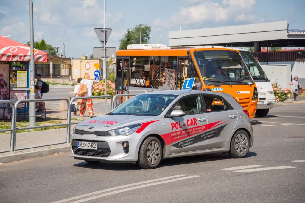 Srebrny samochód z oznaczeniem 'L' i logo 'POL-CAR' na tle pomarańczowego autobusu. Pojazd szkoły jazdy na tle miejskiego krajobrazu w słoneczny dzień.