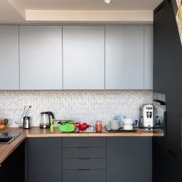 AKIRO - Marcin Abramowicz - Nowoczesna kuchnia z szarymi szafkami górnymi i czarnymi dolnymi, drewniany blat, mozaika na ścianie. Elementy skośne pod schodami. Widoczne tablice rejestracyjne na lodówce.
