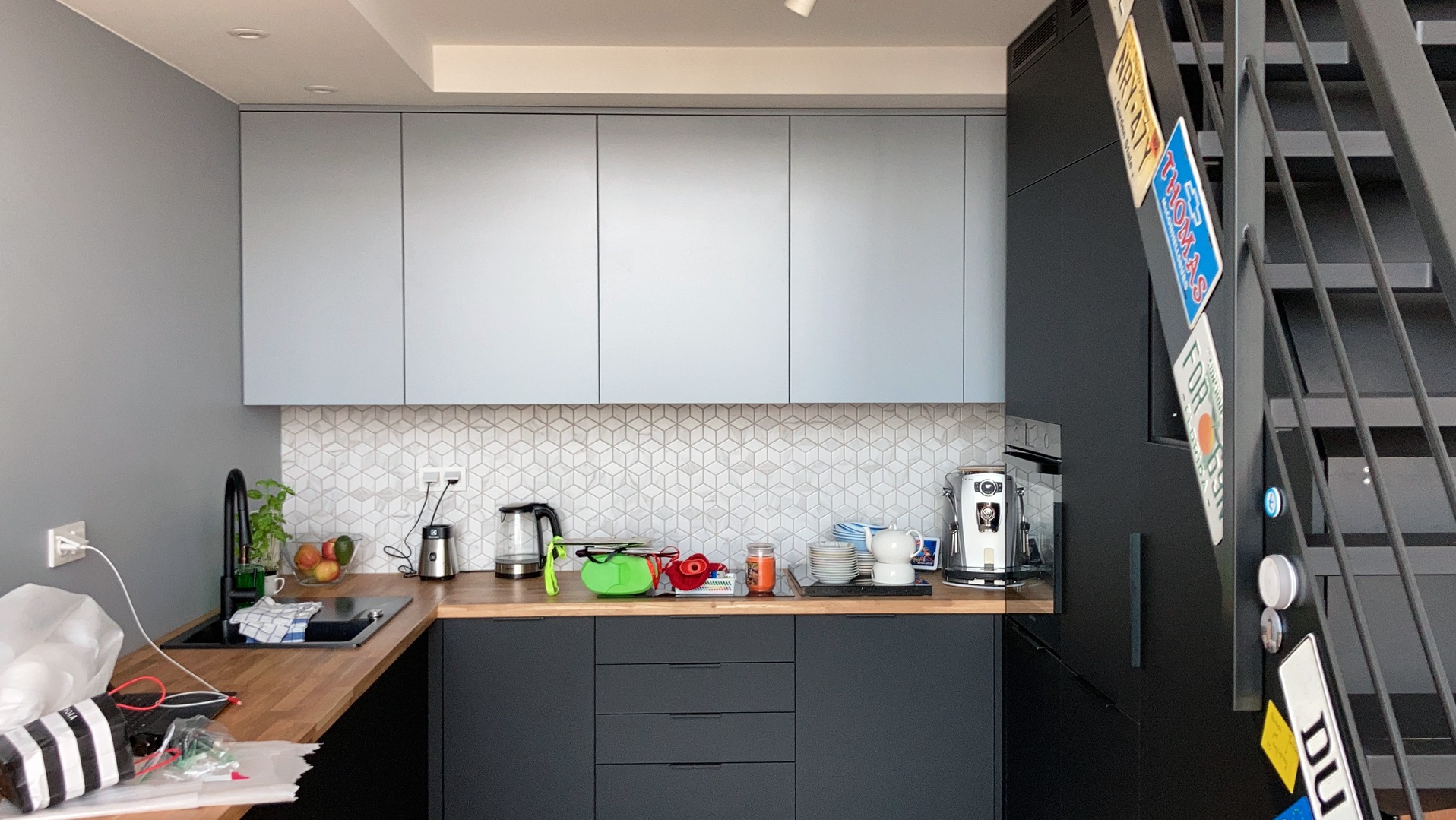 Nowoczesna kuchnia z szarymi szafkami górnymi i czarnymi dolnymi, drewniany blat, mozaika na ścianie. Elementy skośne pod schodami. Widoczne tablice rejestracyjne na lodówce.