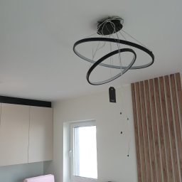 Serhii Novytskyi - Nowoczesna, okrągła lampa sufitowa z trzema pierścieniami w trakcie montażu, widoczne kable i etykieta. W tle drewniana dekoracja ścienna i fragment kuchni.