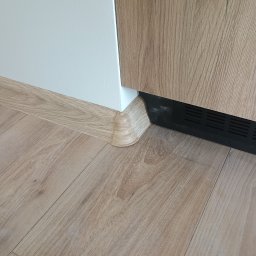 Serhii Novytskyi - Wykończenie podłogi laminowanej z listwą przypodłogową w kolorze drewna, dopasowaną do paneli. Widoczny fragment szafki z drewnianym frontem i biała ściana.