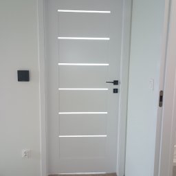 Serhii Novytskyi - Białe drzwi wewnętrzne z poziomymi przeszkleniami i czarną klamką, zamontowane w świeżo pomalowanym na biało pomieszczeniu. Widoczne gniazdko i ślady po montażu.