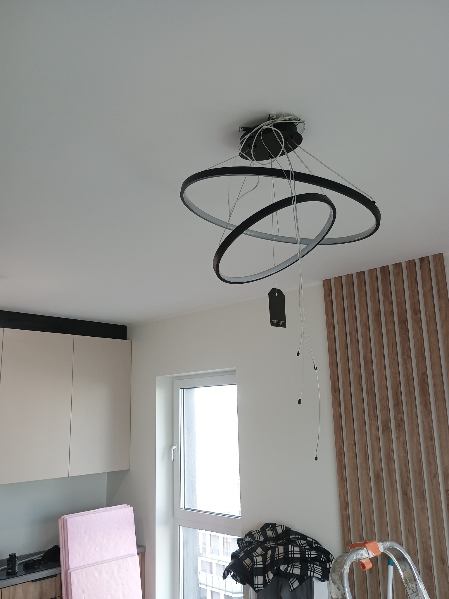 Nowoczesna, okrągła lampa sufitowa z trzema pierścieniami w trakcie montażu, widoczne kable i etykieta. W tle drewniana dekoracja ścienna i fragment kuchni.