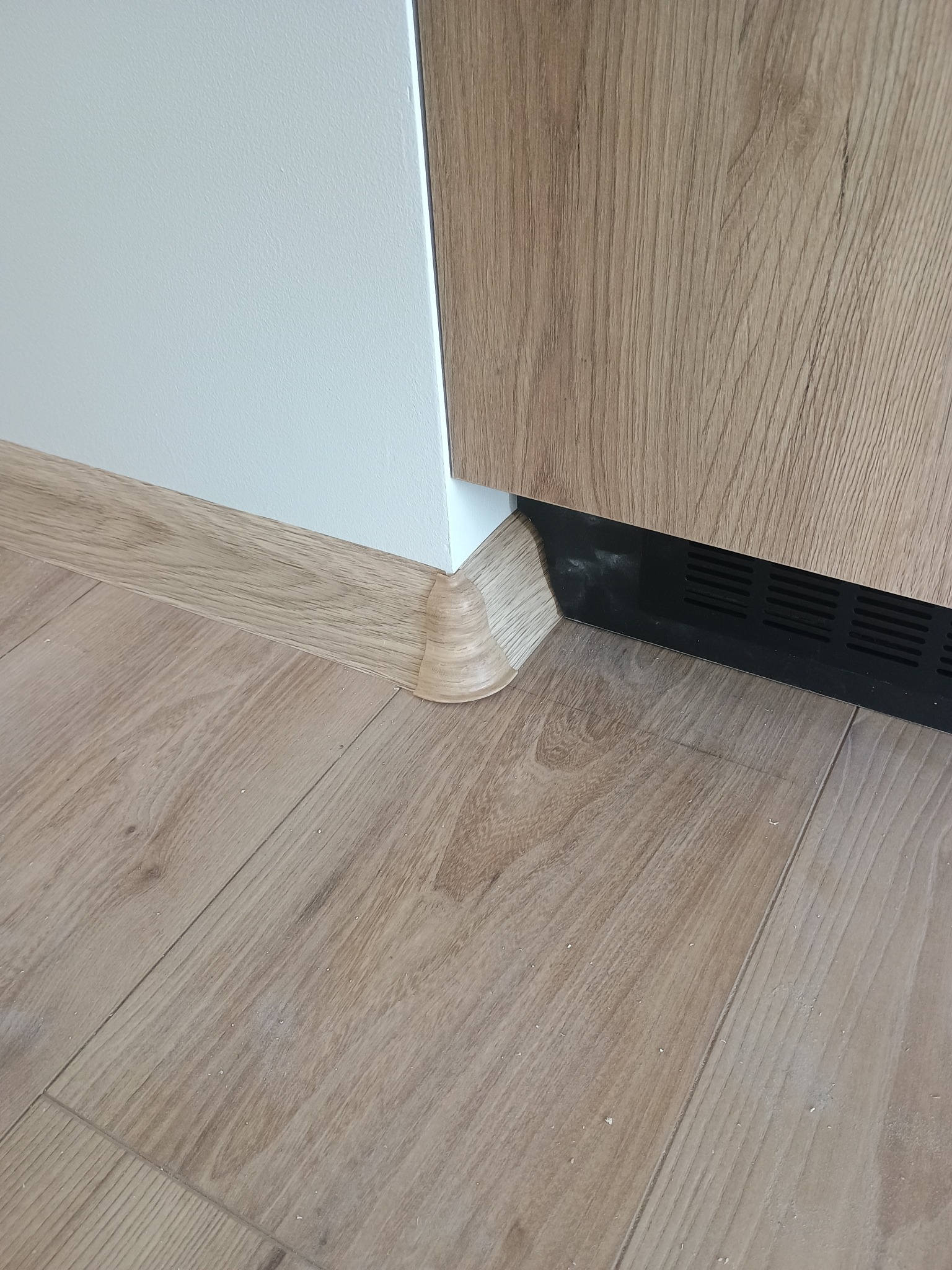 Wykończenie podłogi laminowanej z listwą przypodłogową w kolorze drewna, dopasowaną do paneli. Widoczny fragment szafki z drewnianym frontem i biała ściana.
