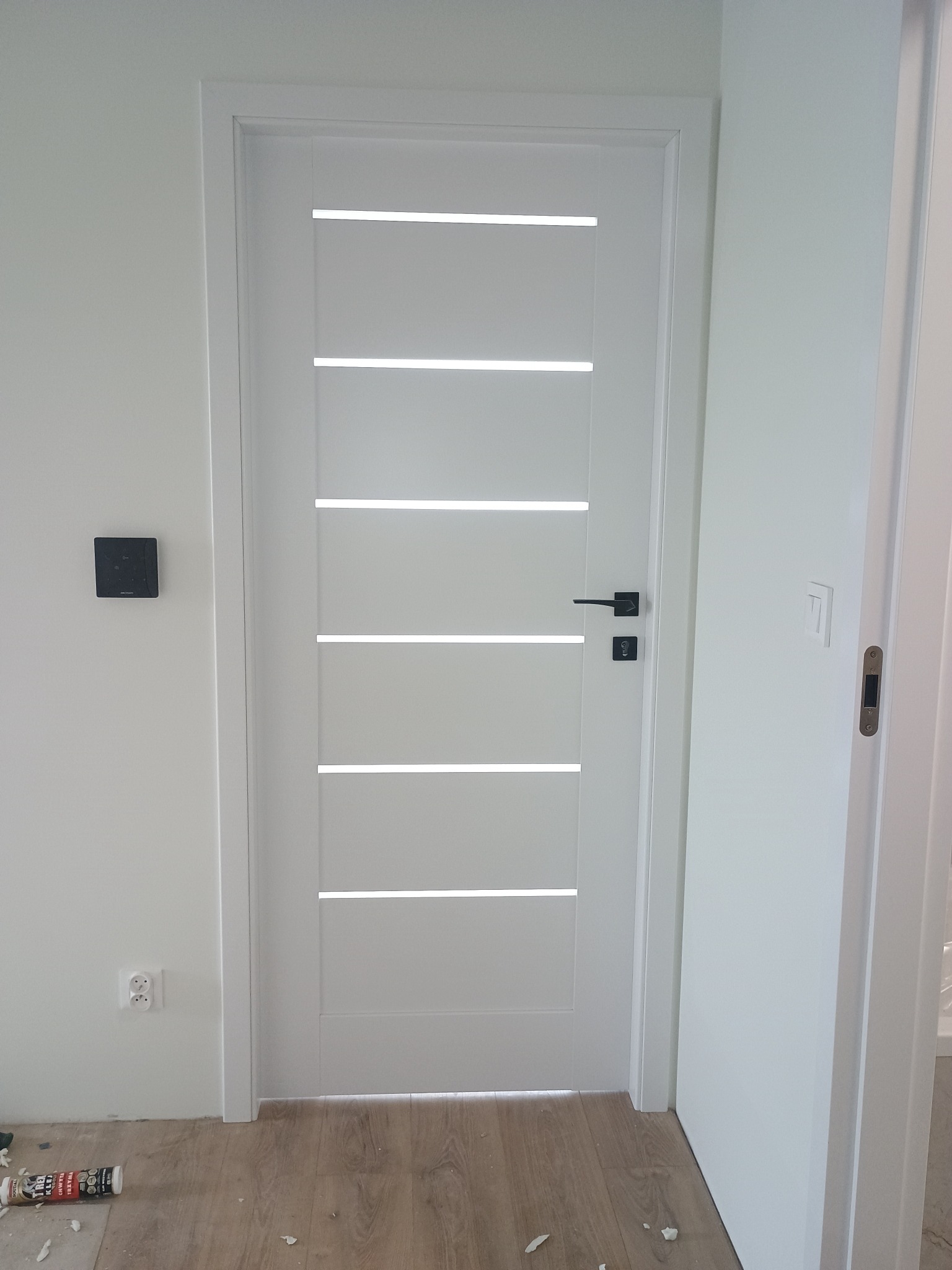 Białe drzwi wewnętrzne z poziomymi przeszkleniami i czarną klamką, zamontowane w świeżo pomalowanym na biało pomieszczeniu. Widoczne gniazdko i ślady po montażu.