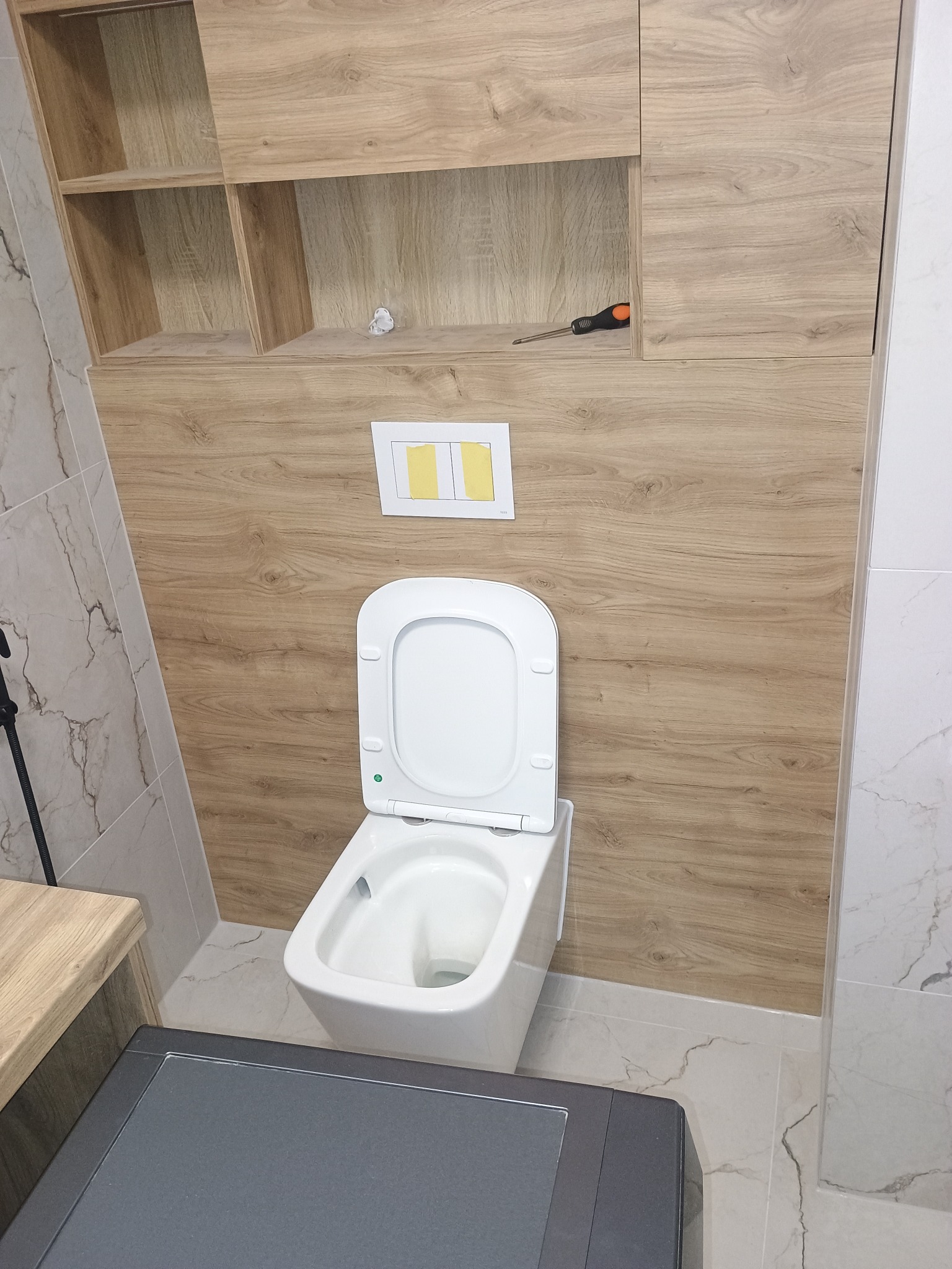 Nowoczesna toaleta podwieszana z deską wolnoopadającą, zamontowana na ścianie z drewnopodobną zabudową i szafkami. Widoczny fragment blatu i marmurowe płytki.