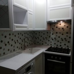 VLADEKBUD - Biała zabudowa kuchenna z szafkami górnymi i dolnymi, zintegrowana z pralką. Uwagę przykuwają heksagonalne płytki na ścianie nad blatem i oświetlenie podszafkowe.