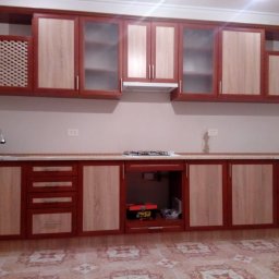 VLADEKBUD - Kompletna zabudowa kuchenna z dwukolorowymi frontami (drewno i bordo) i przeszklonymi szafkami wiszącymi. Widoczny zlew, płyta gazowa i wzorzysta podłoga.