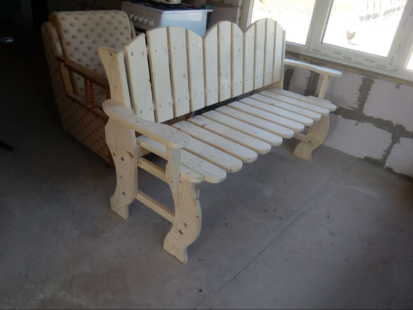 Surowa, drewniana ławka ogrodowa z dekoracyjnymi nogami, ustawiona na betonowej posadzce, z widocznym oknem i fragmentem ceglanej ściany w tle.