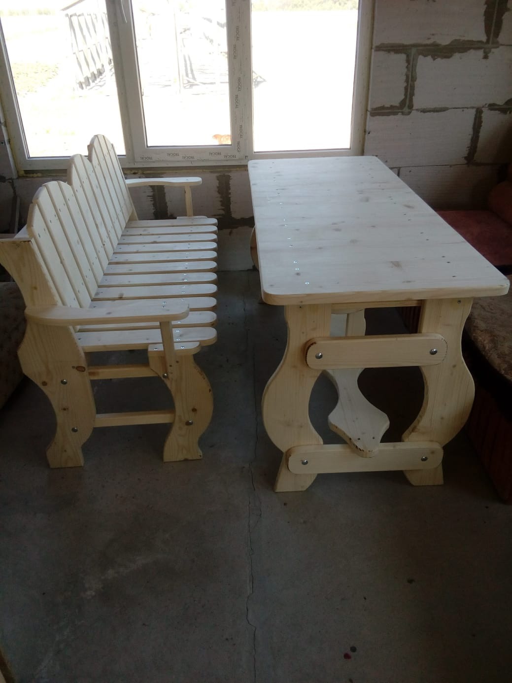 Drewniany stół i ławka ogrodowa z jasnego drewna, widoczne śruby mocujące, surowe wykończenie, tło z pustaków.