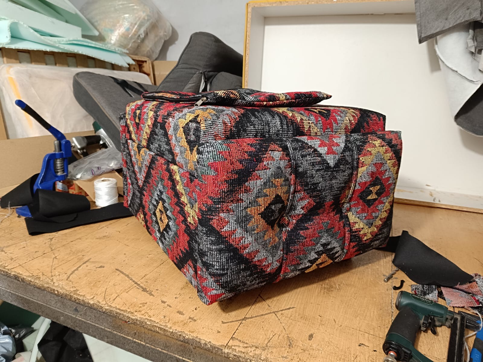 Tapicerowany puf z wzorzystej tkaniny w odcieniach czerwieni, czerni i żółci, leżący na drewnianym stole w warsztacie tapicerskim. Obok narzędzia i materiały.