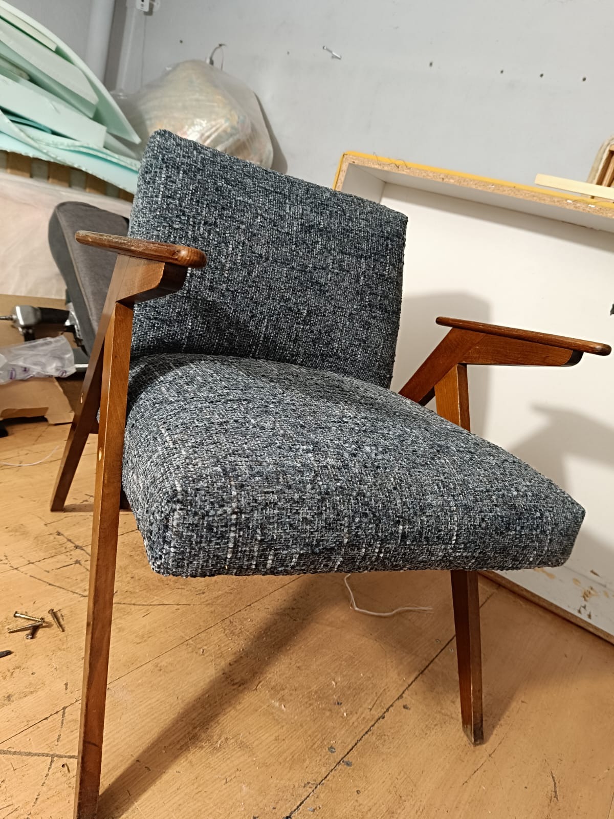 Odrestaurowany fotel z drewnianymi podłokietnikami i szarą tapicerką o wyraźnej fakturze, stojący w warsztacie. Widoczne ślady renowacji i narzędzia w tle.