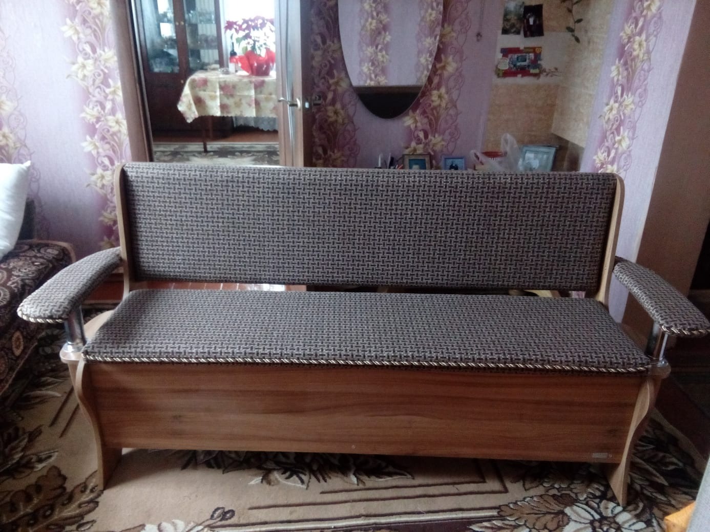 Tapicerowana ławka z drewnianą skrzynią w stylu retro, na tle wzorzystej tapety i dywanu. Widoczne odbicie w lustrze i fragment wnętrza pokoju.