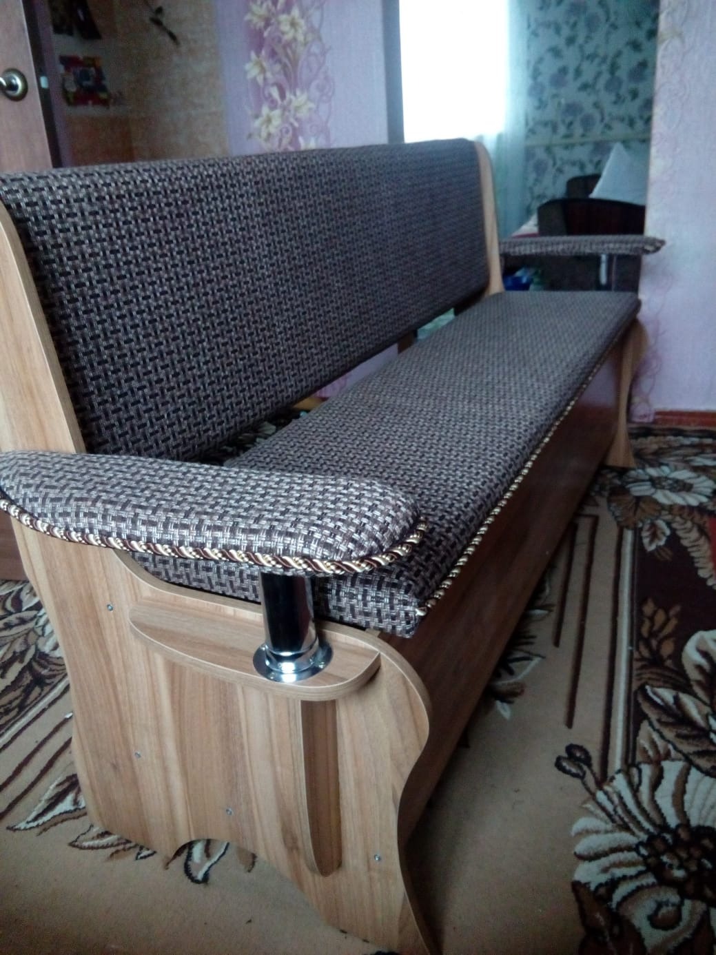 Tapicerowana ławka z drewnianymi elementami, wzorzysta tkanina obiciowa w odcieniach brązu, widok z boku, na tle wzorzystej tapety i dywanu.