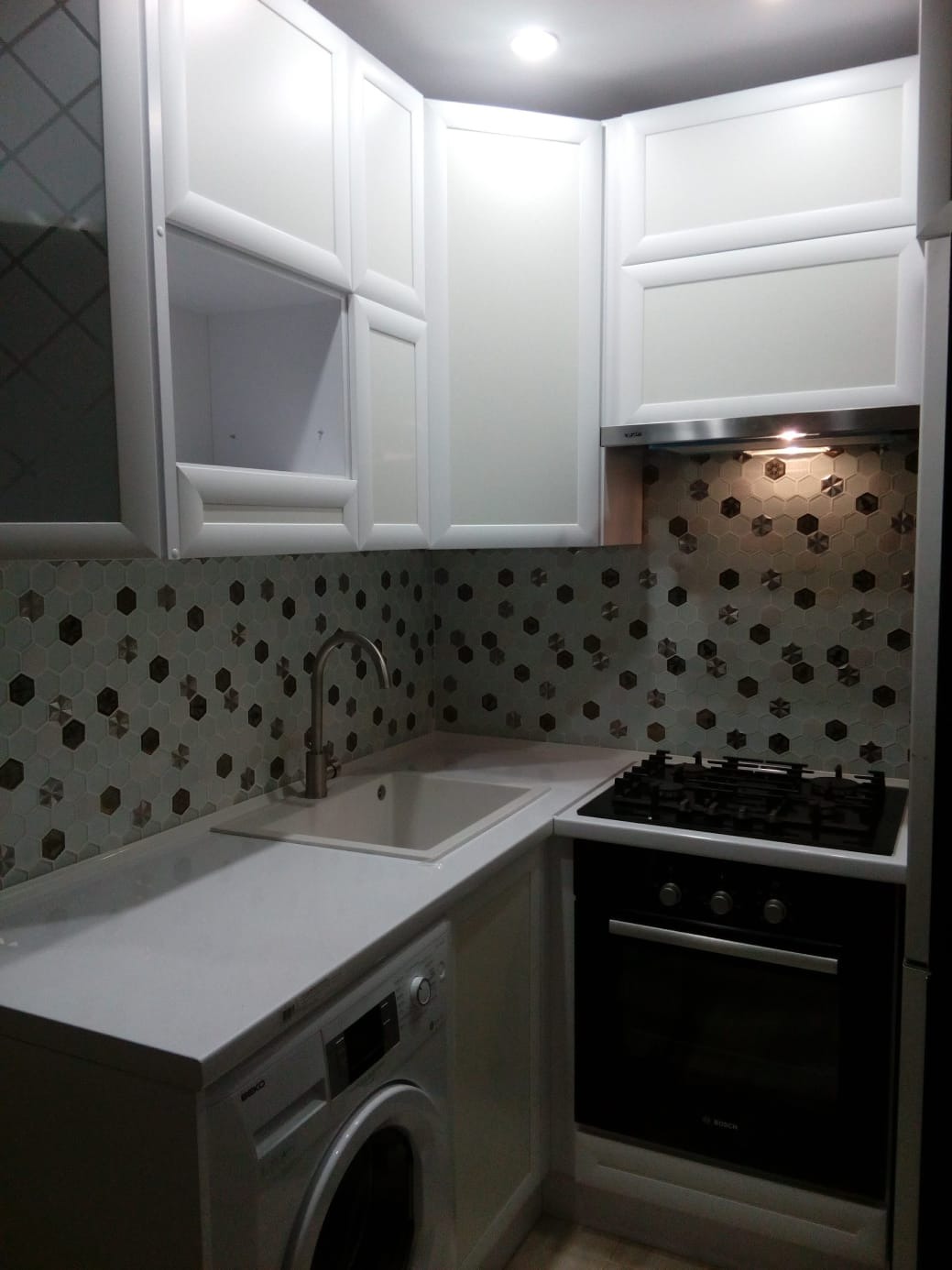 Biała zabudowa kuchenna z szafkami górnymi i dolnymi, zintegrowana z pralką. Uwagę przykuwają heksagonalne płytki na ścianie nad blatem i oświetlenie podszafkowe.