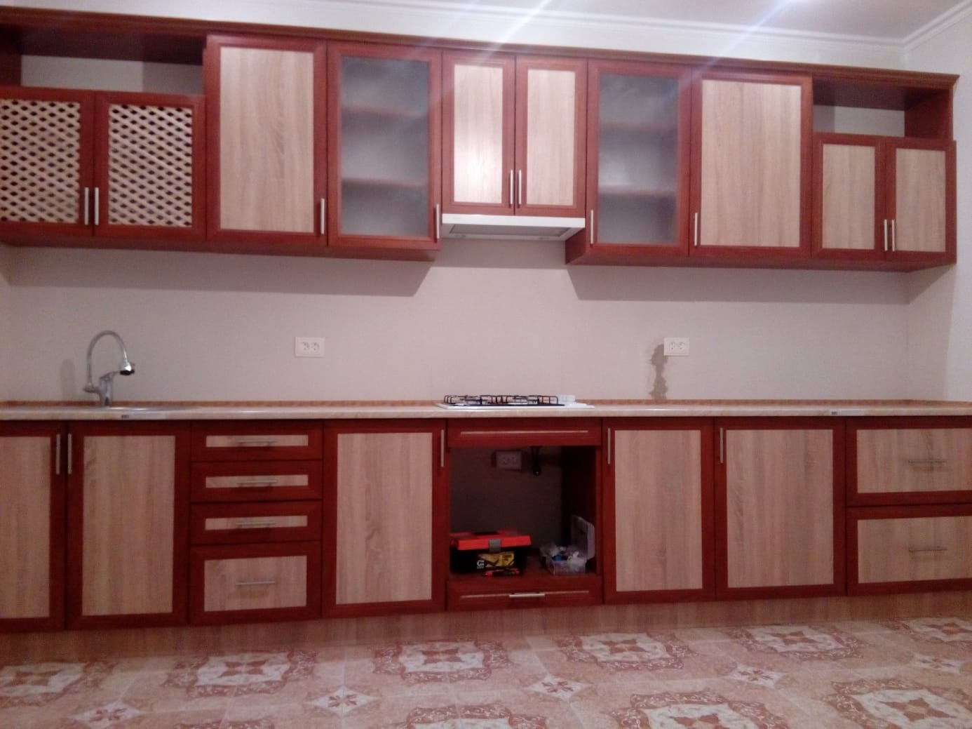 Kompletna zabudowa kuchenna z dwukolorowymi frontami (drewno i bordo) i przeszklonymi szafkami wiszącymi. Widoczny zlew, płyta gazowa i wzorzysta podłoga.