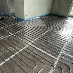 FESTGATE SPÓŁKA Z OGRANICZONĄ ODPOWIEDZIALNOŚCIĄ - Szare rury ogrzewania podłogowego ułożone na folii z czerwoną siatką w surowym wnętrzu. Widoczne fragmenty ścian i okno. Instalacja przed wylaniem posadzki.