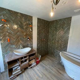 FESTGATE SPÓŁKA Z OGRANICZONĄ ODPOWIEDZIALNOŚCIĄ - Łazienka w trakcie wykańczania: ułożona glazura w jodełkę na ścianach, drewnopodobna podłoga, wolnostojąca wanna w folii, szafka pod umywalkę z otwartymi szufladami.