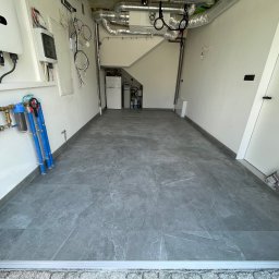 FestGate Firma Budowlana - Szare płytki podłogowe w garażu/pomieszczeniu gospodarczym. Widoczne instalacje, rury, filtry wody, elementy elektryczne i wentylacyjne. Minimalistyczne wnętrze.