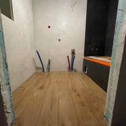 FestGate Firma Budowlana - Wykończona podłoga z paneli drewnopodobnych w łazience z częściowo położonymi płytkami ściennymi i instalacją wodną. Widoczne niedokończone obramowanie drzwi.