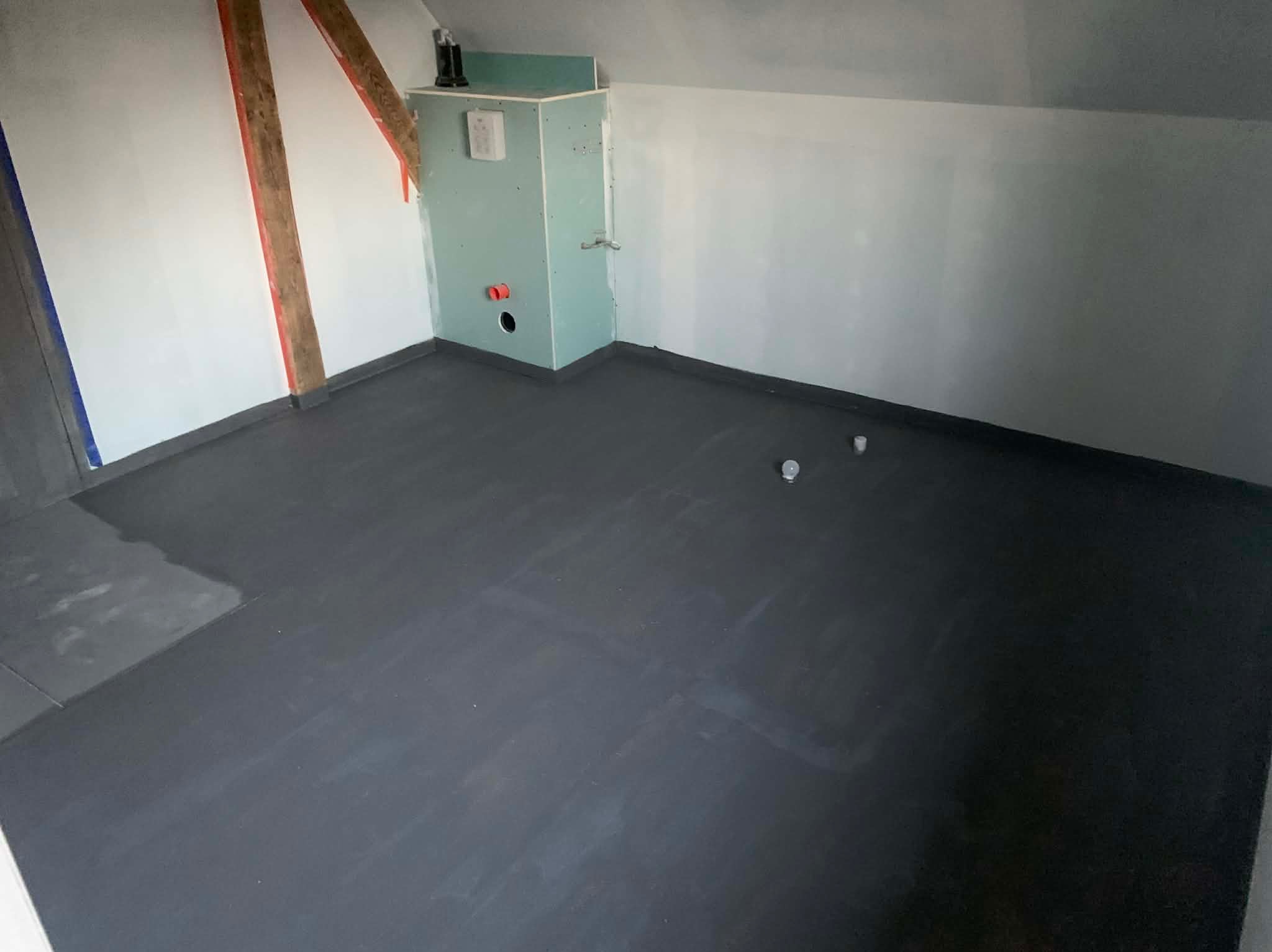 Szara wylewka samopoziomująca na poddaszu, przygotowana pod ogrzewanie podłogowe. Widoczna instalacja wodna w ścianie i fragment drewnianej konstrukcji dachu.