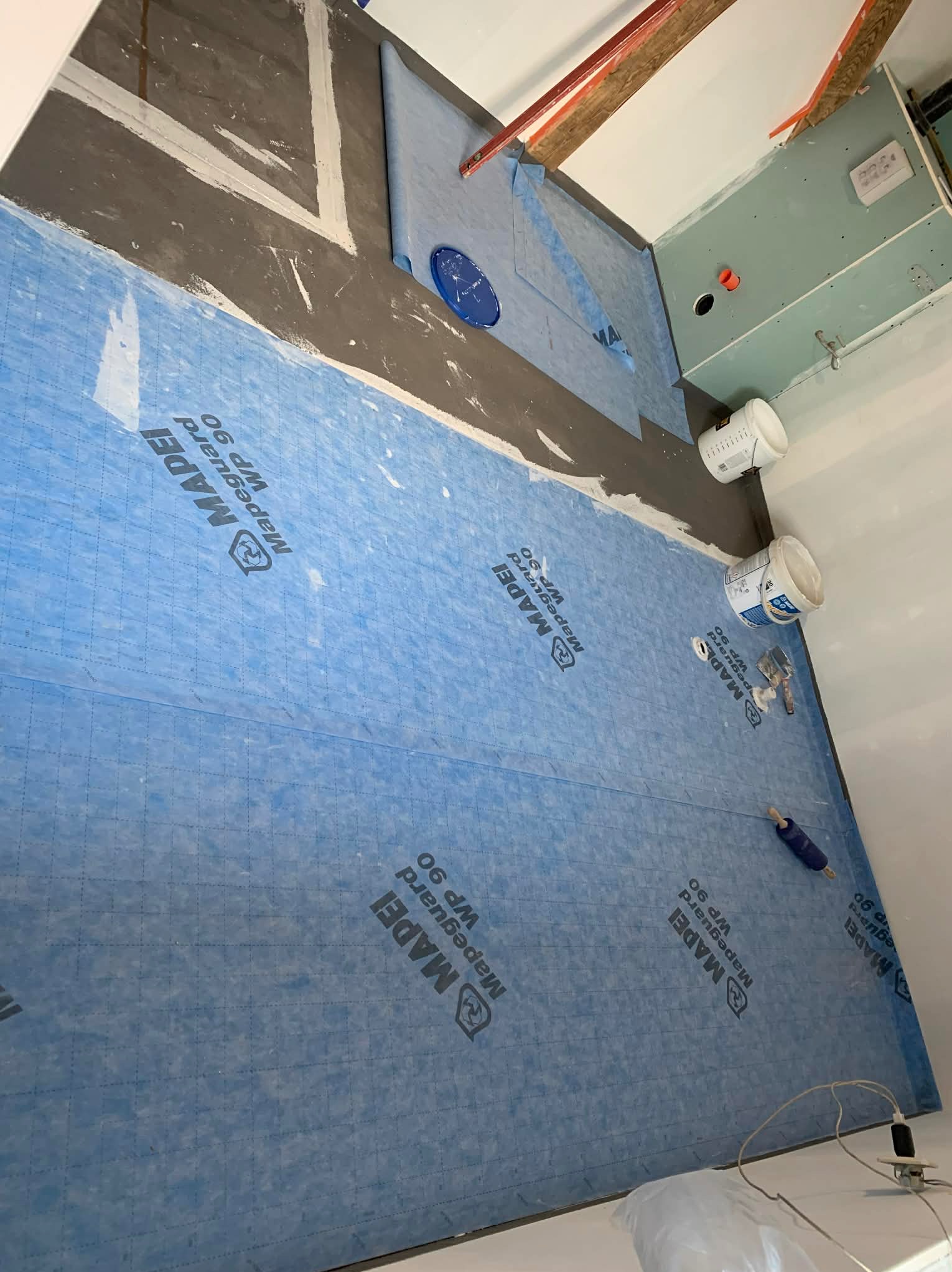 Hydroizolacja podłoża w łazience, niebieska membrana Mapeguard WP 90, przygotowanie do położenia płytek. Widoczne wiaderka z zaprawą i narzędzia glazurnicze.