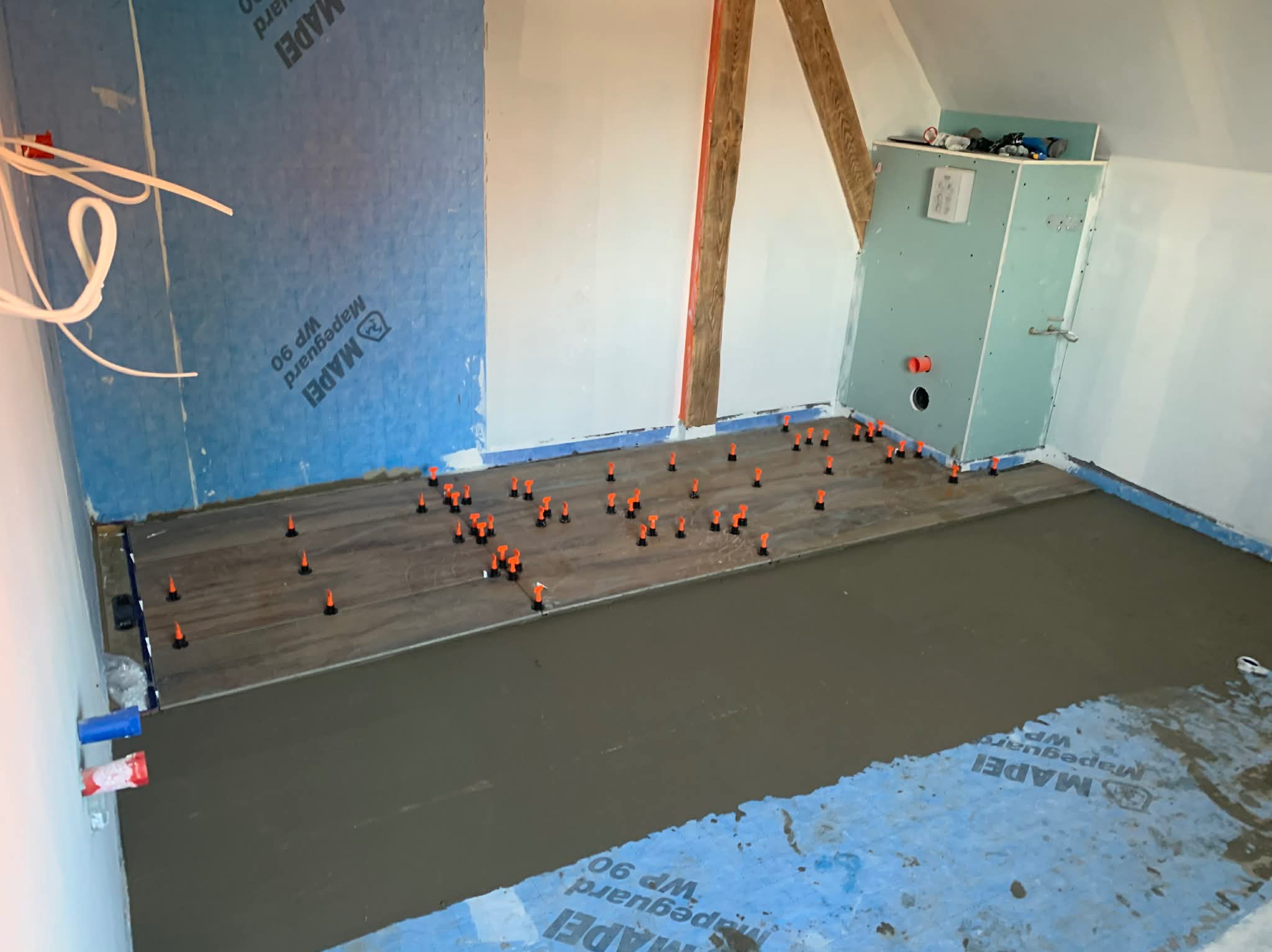 Układanie płytek imitujących drewno w łazience na podkładzie z wylewki samopoziomującej. Widoczne poziomujące klipsy i elementy instalacji hydraulicznej.