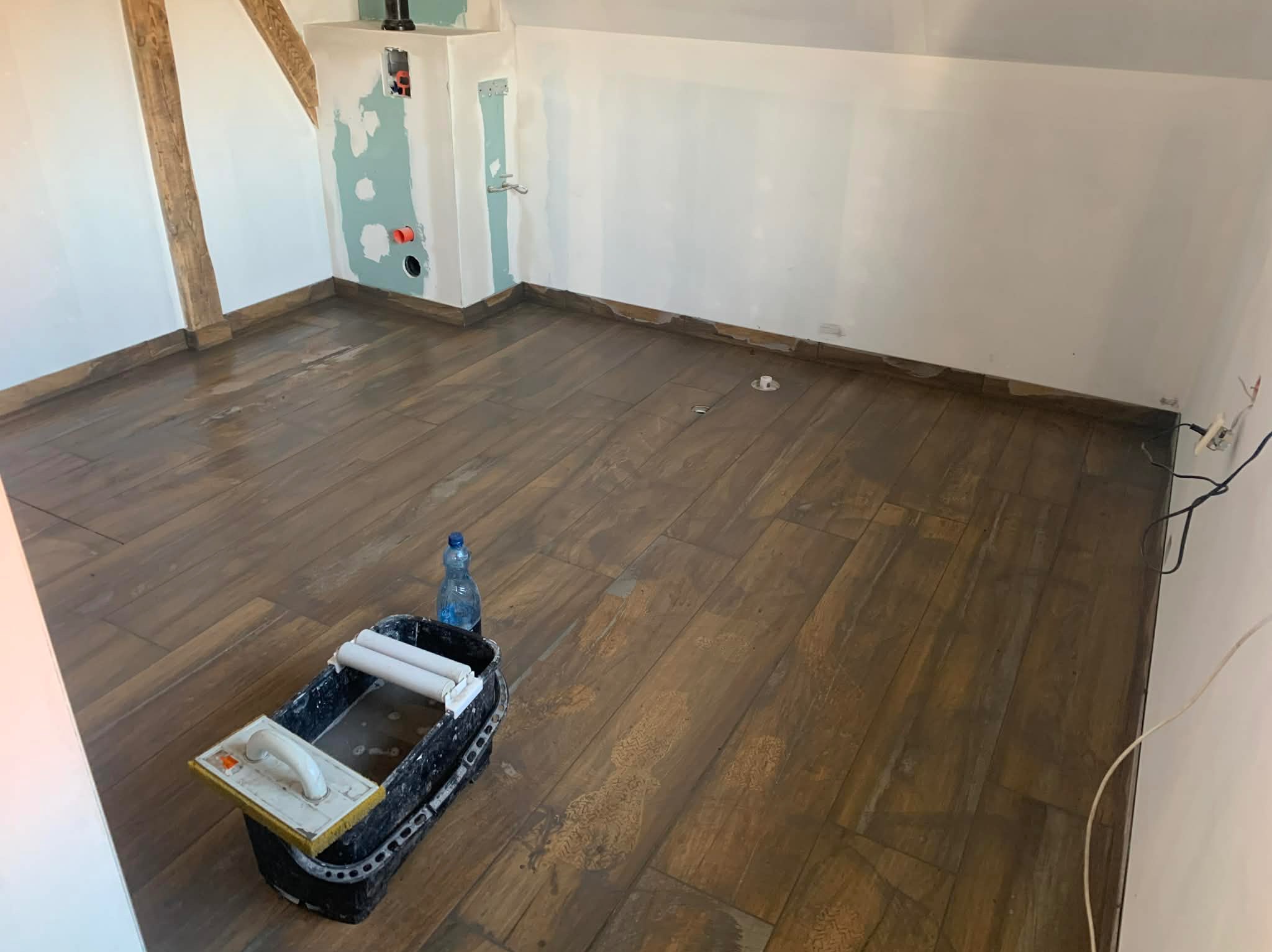 Ułożona podłoga z płytek imitujących drewno w pokoju na poddaszu. Widoczny kubeł glazurniczy i butelka wody. Ściany przygotowane do malowania.