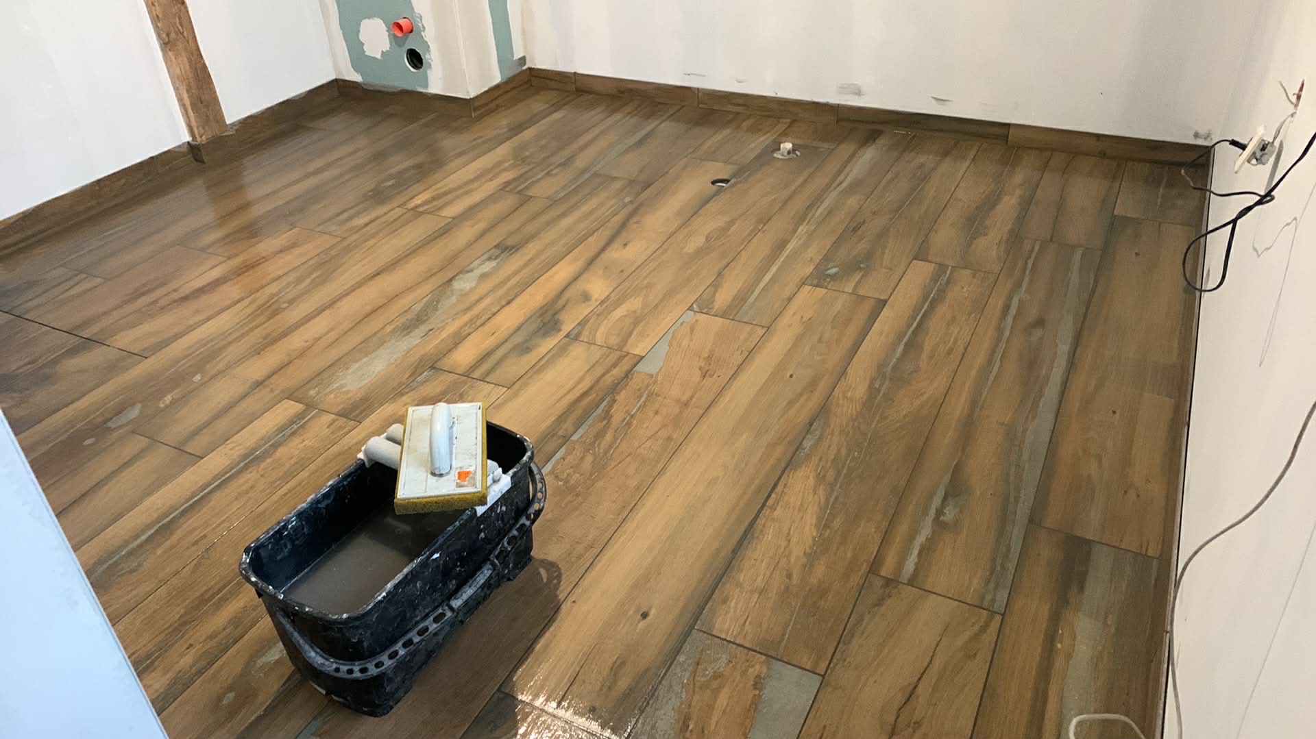 Ułożona podłoga z płytek imitujących drewno w jasnym pomieszczeniu. Widoczny kubeł z narzędziami glazurniczymi. Świeżo położona podłoga, fuga jeszcze mokra.