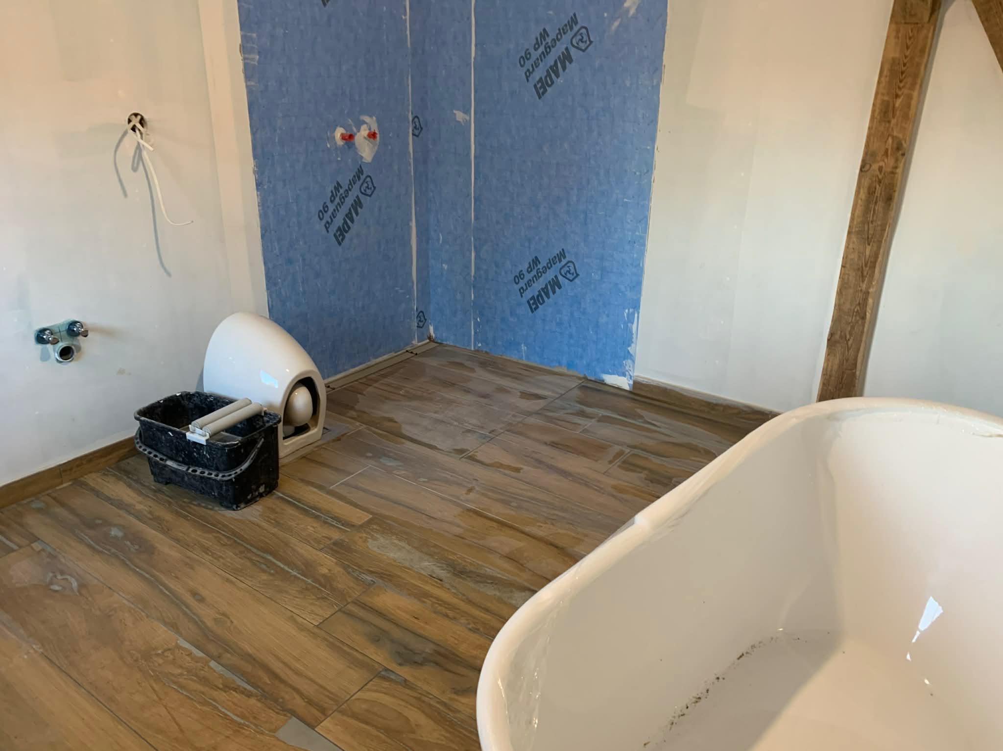 Remont łazienki: ułożona podłoga z płytek imitujących drewno, zabezpieczone ściany folią w płachach, wanna i wiadro z narzędziami. Widoczna drewniana belka stropowa.