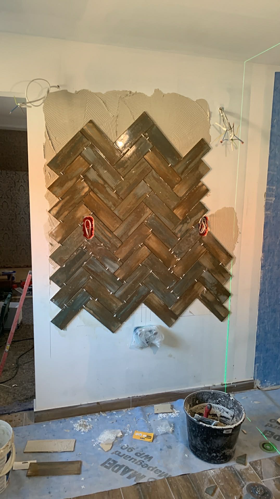 Układanie płytek w jodełkę na ścianie, widoczne narzędzia glazurnicze i laserowy niwelator. Etap prac z częściowo ułożonym wzorem i widoczną zaprawą klejową.