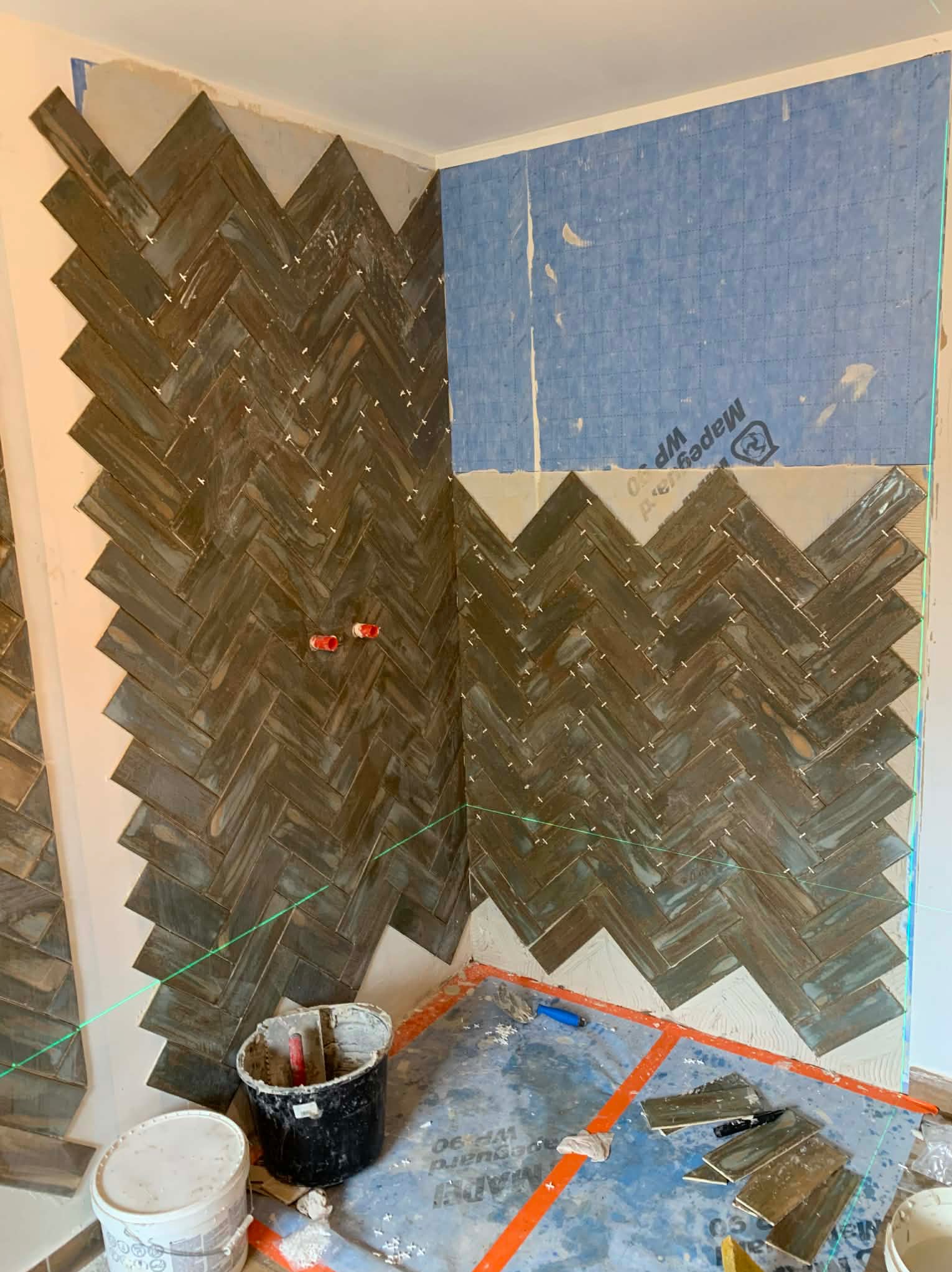 Układanie płytek w jodełkę na ścianie, widoczne narzędzia glazurnicze, laser krzyżowy, fragment ściany zabezpieczony folią. Prace w toku, brązowo-szare płytki.