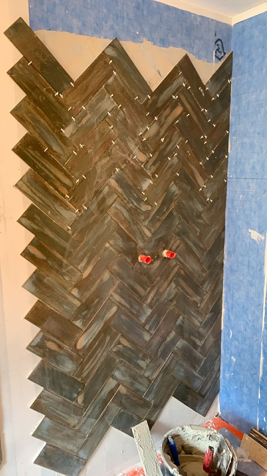 Ściana w trakcie wykańczania z ułożoną glazurą w jodełkę, widoczne krzyżyki dystansowe i fragment instalacji. W tle zabezpieczona ściana i wiadro z zaprawą.