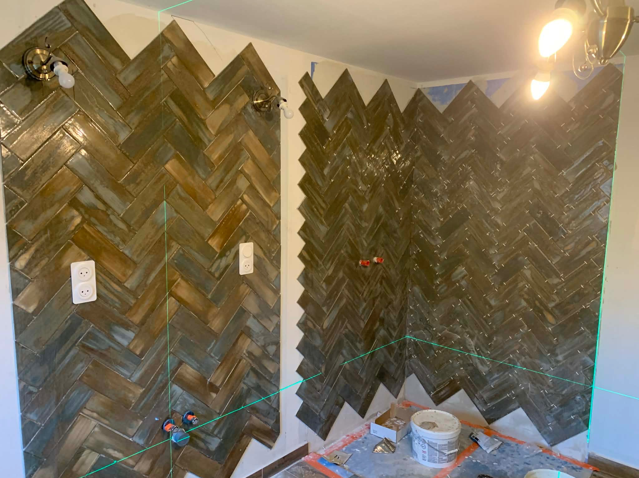 Układanie brązowo-szarych płytek w jodełkę na ścianach łazienki, widoczne gniazdka elektryczne, elementy instalacji i laserowy niwelator. Prace glazurnicze w toku.