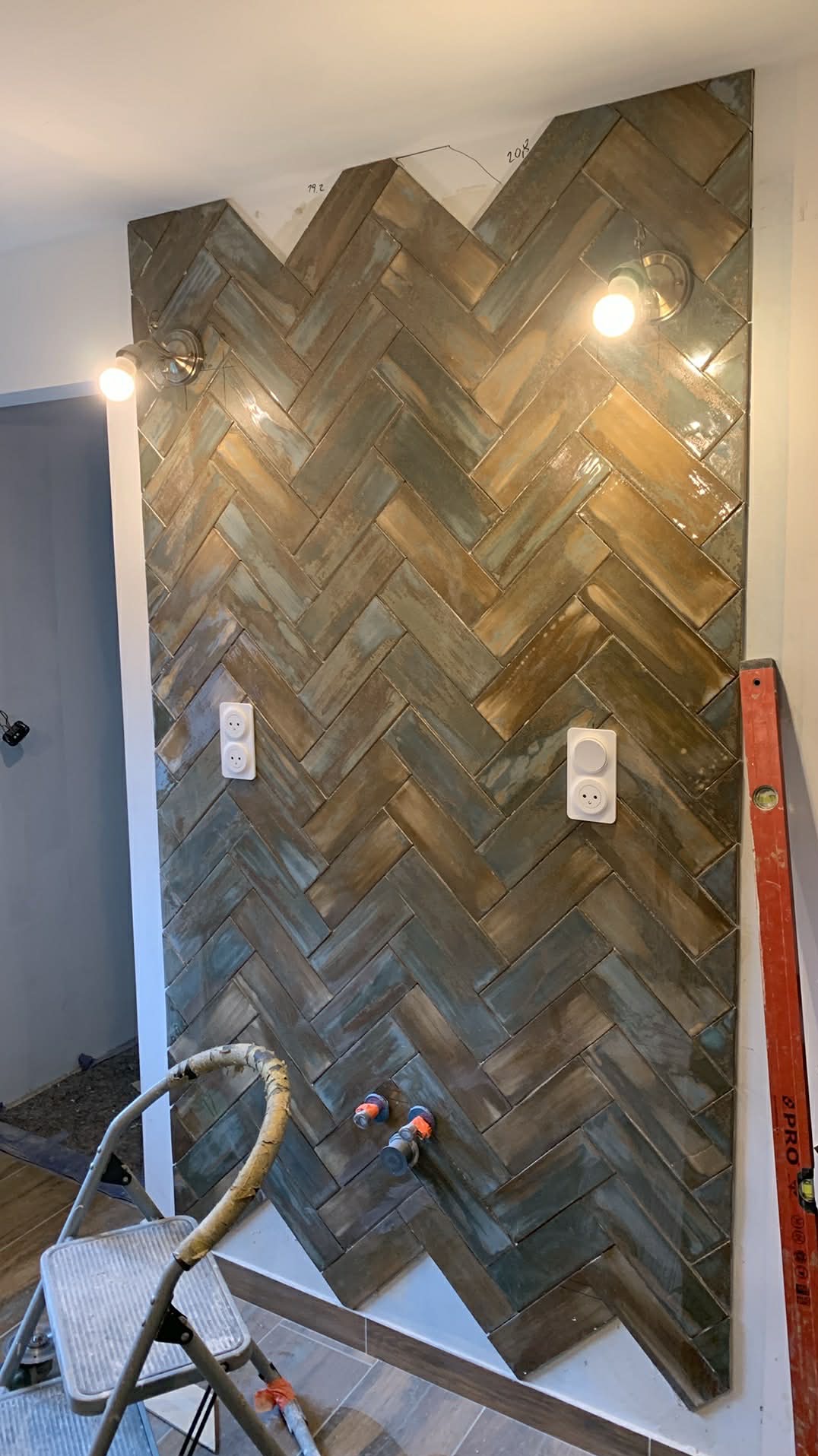 Ściana wyłożona błyszczącymi, brązowo-turkusowymi płytkami w jodełkę, z zamontowanymi gniazdkami i lampami. Widoczna drabina i poziomica.