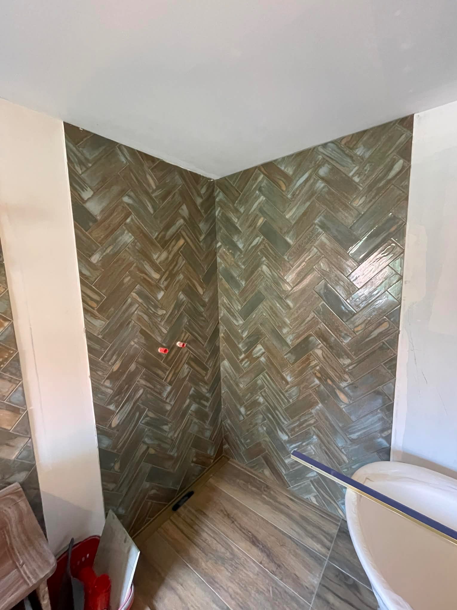 Ukończona ściana w łazience z brązowo-zielonymi płytkami ułożonymi w jodełkę, widoczne fragmenty wanny i podłogi z płytek imitujących drewno.