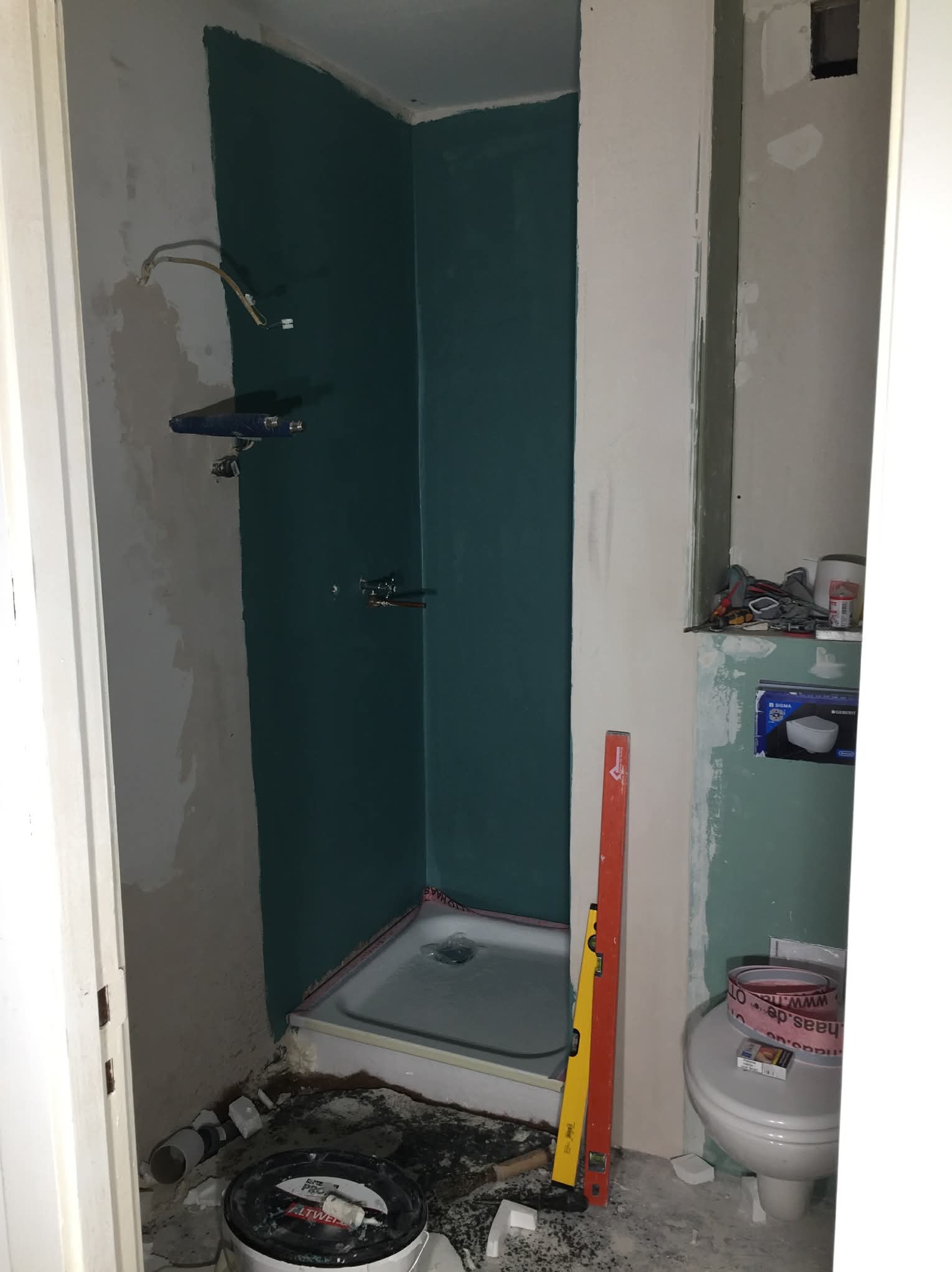 Niedokończona łazienka z brodzikiem, przygotowana do położenia płytek. Widoczne narzędzia, poziomice i instalacja wodna. Ściany częściowo pomalowane na zielono.