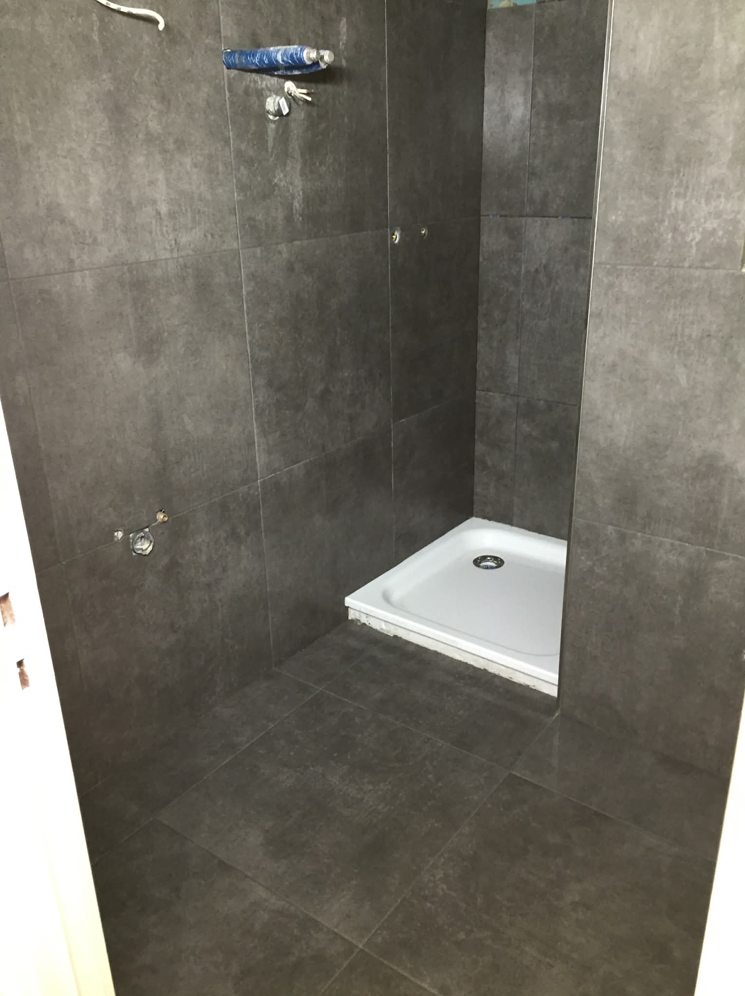 Wykończona kabina prysznicowa z ciemnymi płytkami, widoczne przyłącza hydrauliczne i biały brodzik. Minimalistyczny design, surowe wykończenie.