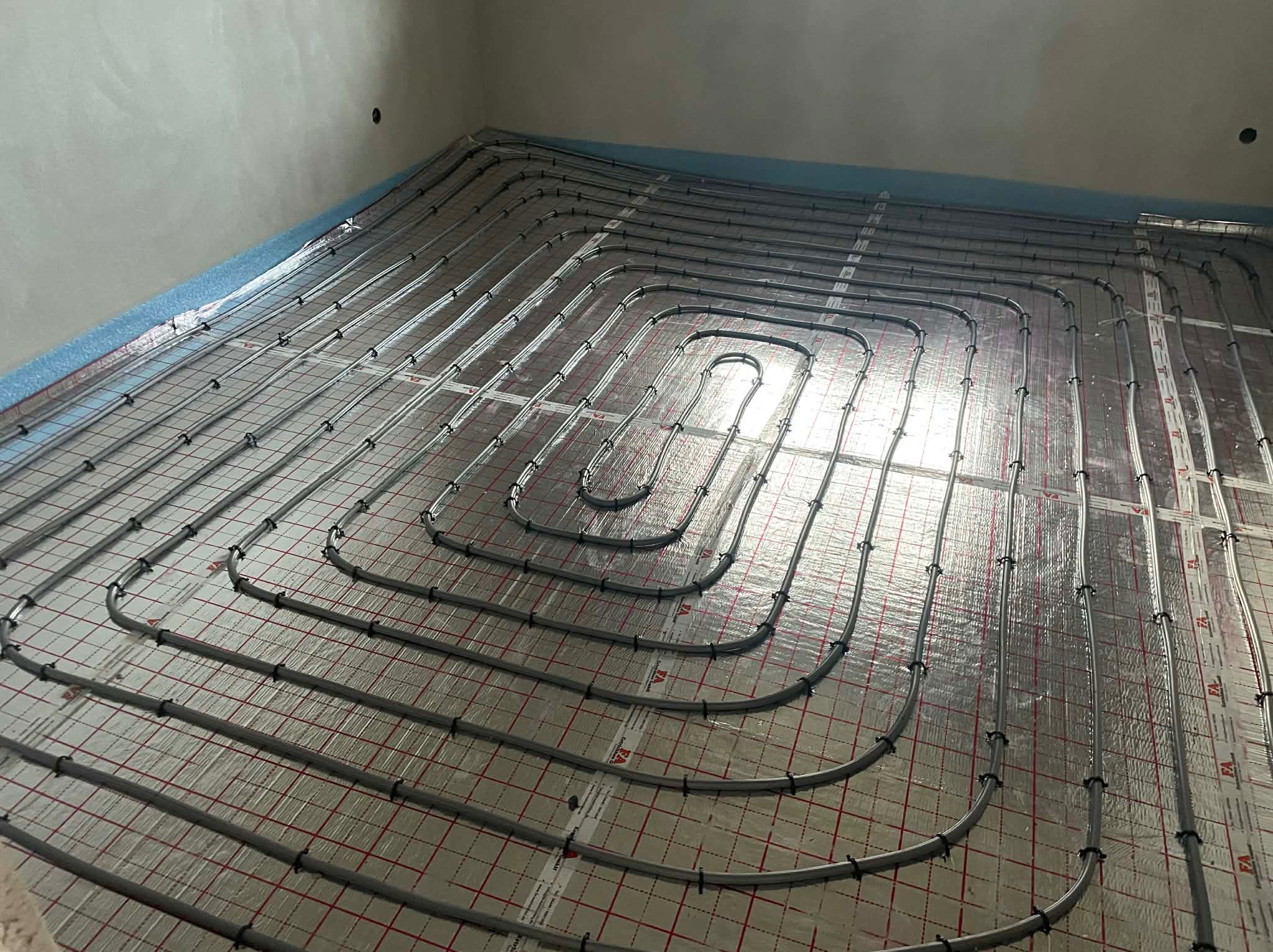 Szare rury ogrzewania podłogowego ułożone spiralnie na srebrnej folii z czerwoną siatką, widok z góry, surowy stan wykończenia wnętrza.