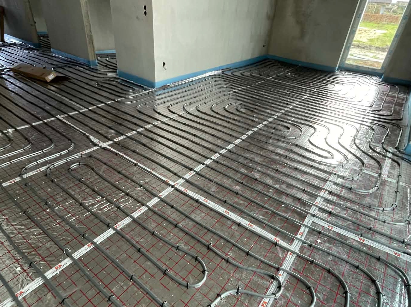 Szare rury ogrzewania podłogowego ułożone na folii z czerwoną siatką w surowym wnętrzu. Widoczne fragmenty ścian i okno. Instalacja przed wylaniem posadzki.