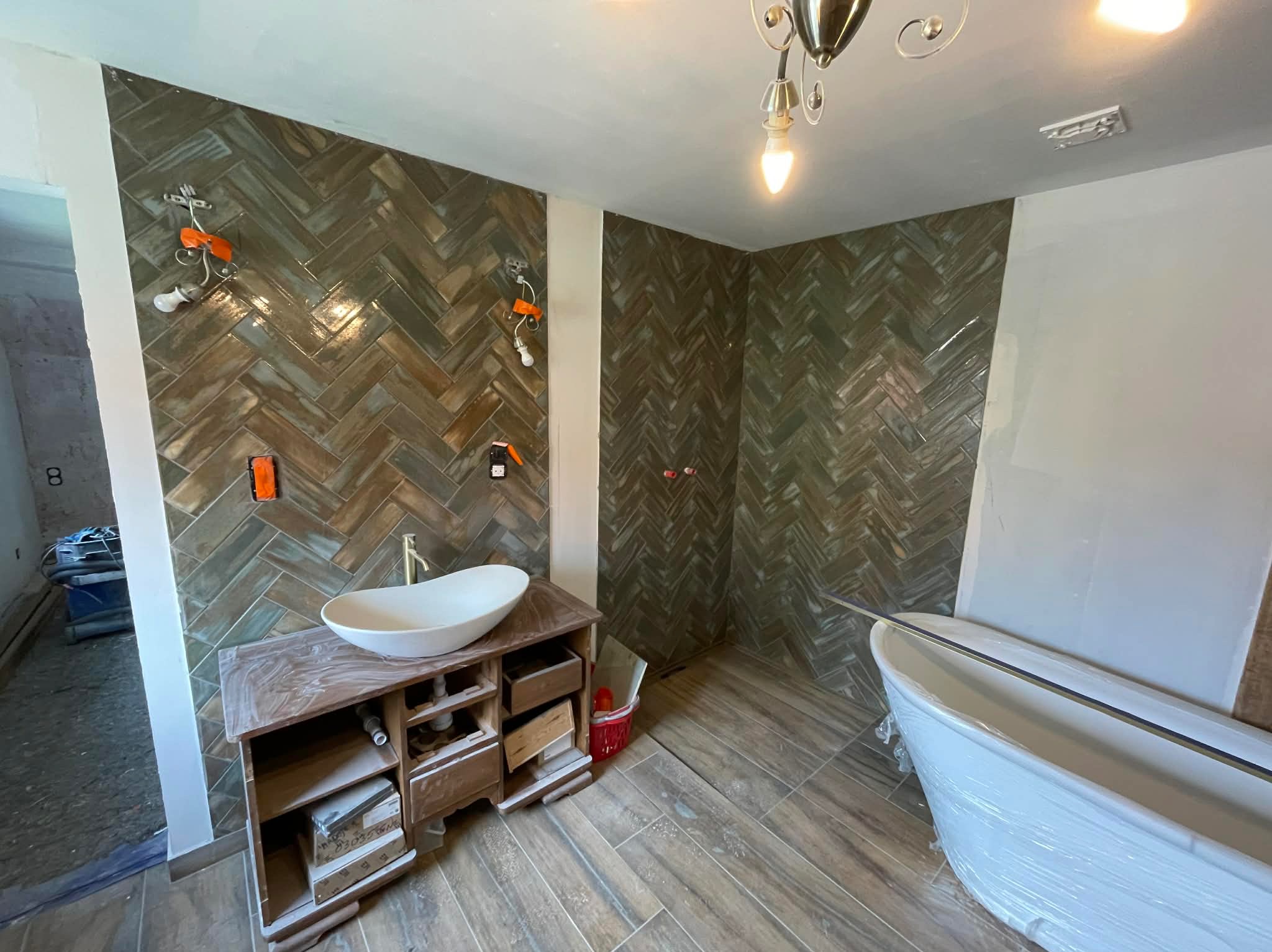 Łazienka w trakcie wykańczania: ułożona glazura w jodełkę na ścianach, drewnopodobna podłoga, wolnostojąca wanna w folii, szafka pod umywalkę z otwartymi szufladami.