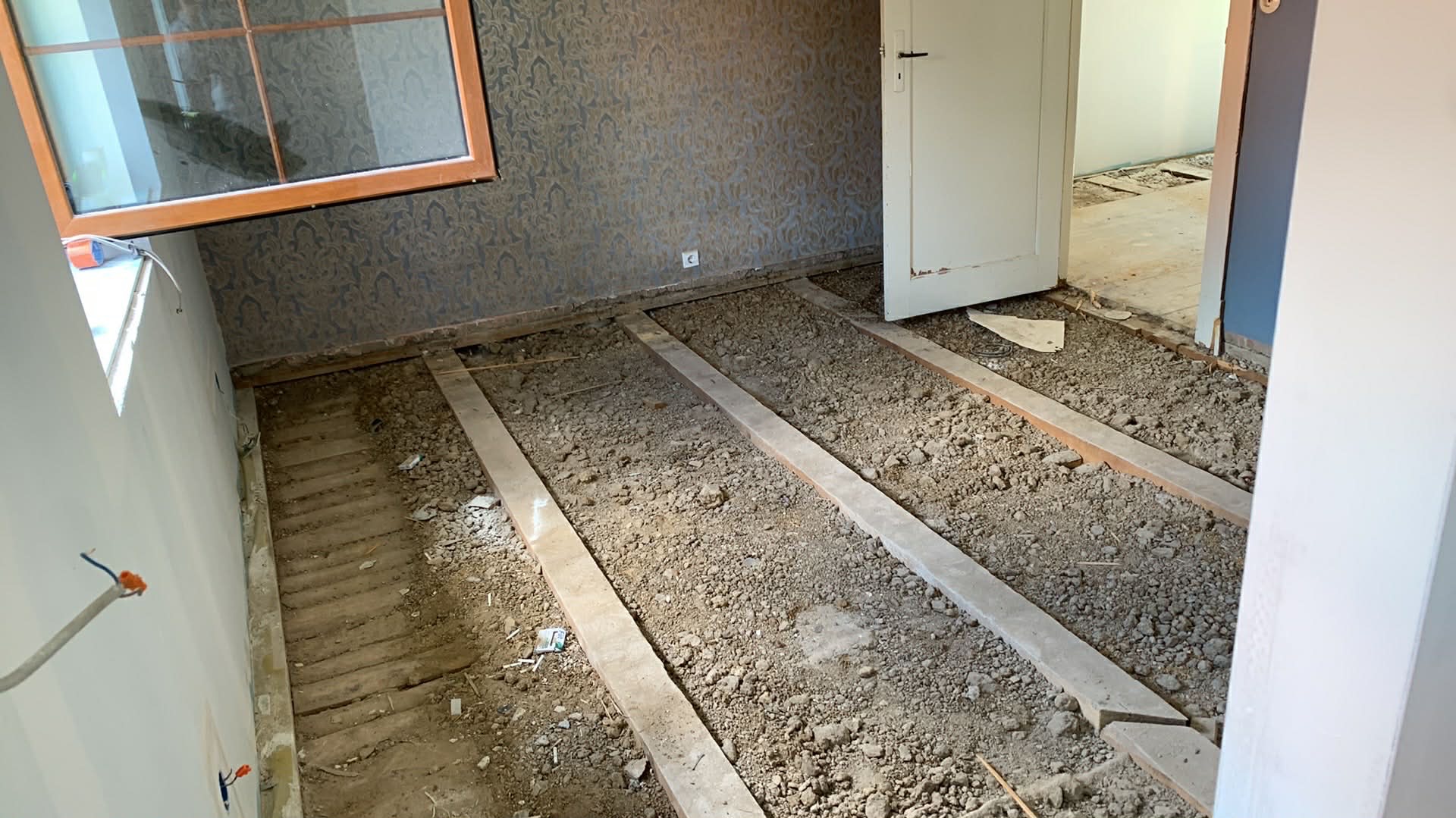 Przygotowanie podłoża pod ogrzewanie podłogowe: widoczne listwy i gruz na podłodze w remontowanym pokoju z oknem i tapetą w tle. Otwarta biała futryna drzwi.