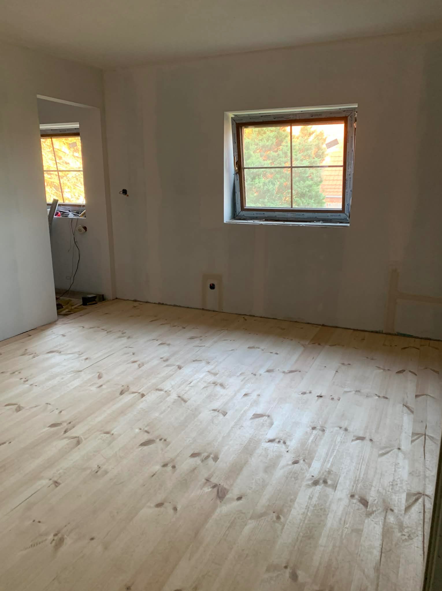 Jasne, drewniane deski podłogowe w pokoju z oknami. Ściany białe, przygotowane do malowania. Widoczne gniazdka elektryczne i fragment instalacji.