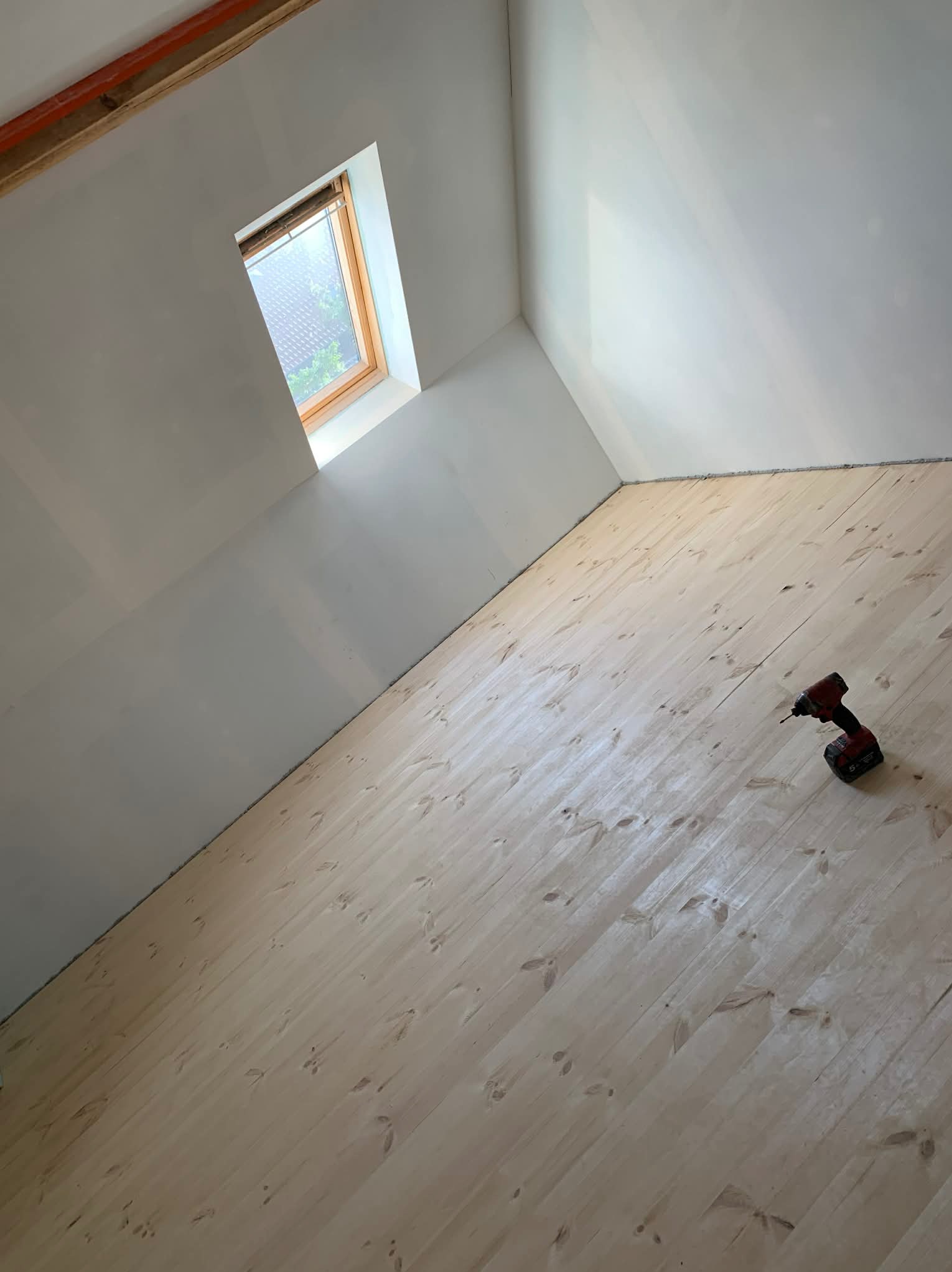 Podłoga z jasnego drewna na poddaszu, widoczne okno dachowe i wkrętarka akumulatorowa. Ukończony etap prac wykończeniowych, gotowy do dalszych działań.