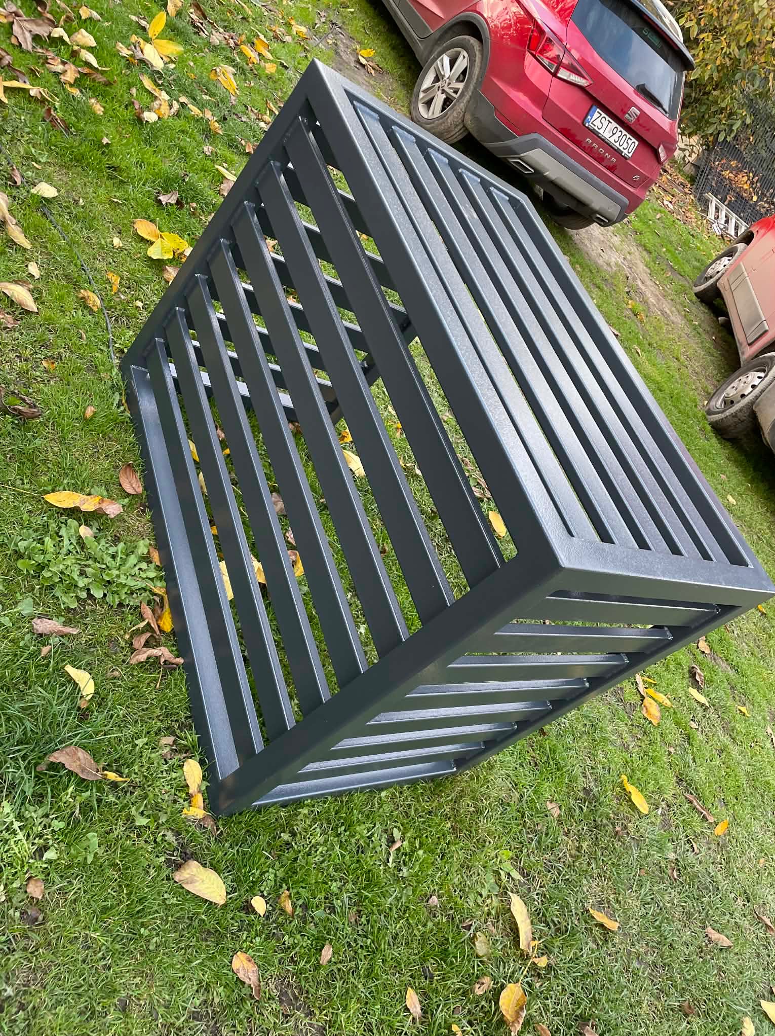 Szary, metalowy fragment ogrodzenia panelowego leży na trawie z opadłymi liśćmi, w tle zaparkowane samochody. Widok z góry, ukośny kadr.