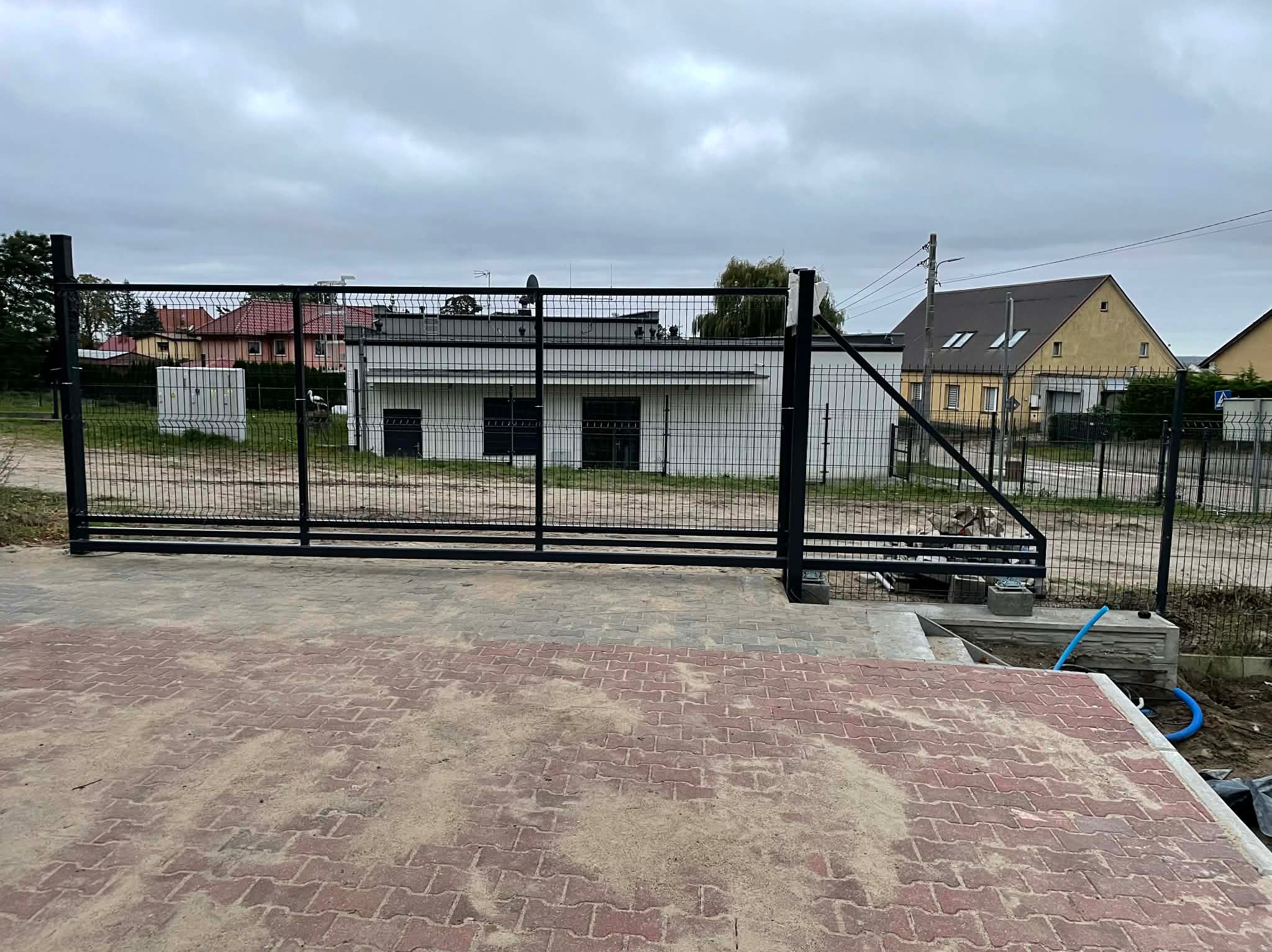 Czarna, przesuwna brama z siatki na posesji z kostką brukową. Widoczne budynki w tle i pochmurne niebo. Brama częściowo otwarta, z widocznymi elementami montażowymi.