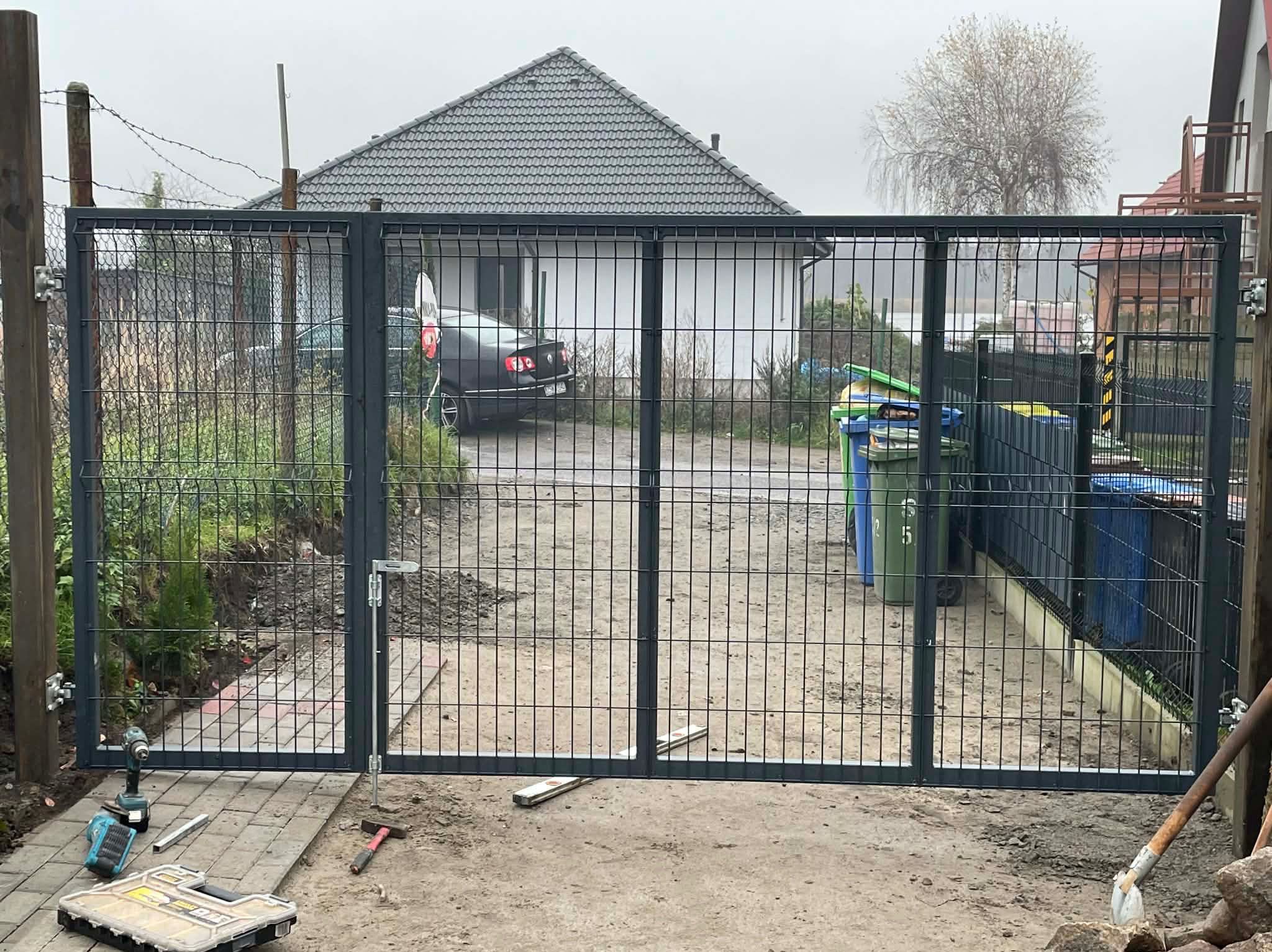Nowoczesna, metalowa brama panelowa w trakcie montażu. Widoczne narzędzia, poziomica i skrzynka narzędziowa. W tle dom jednorodzinny i drzewo bez liści.
